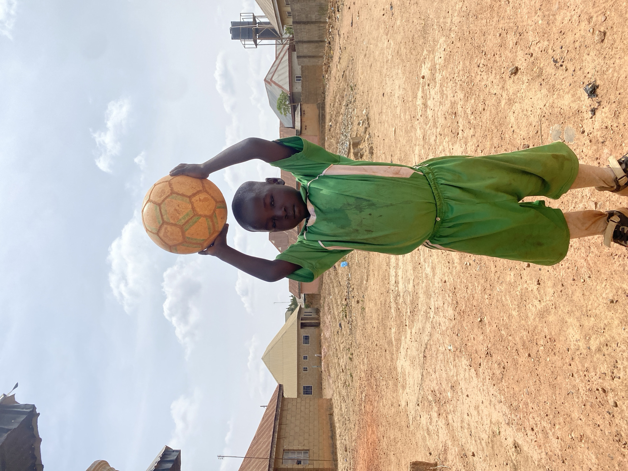An image taken in Rido, Nigeria, depicts a young boy standing centrally in an open, unpaved area of reddish-brown earth under a partially cloudy sky. The boy, dressed in a green t-shirt with white side stripes and matching green shorts, holds an orange football with green panel outlines above his head with both hands, looking directly at the camera. He wears brown shoes with light-colored straps. The ground is dry and dusty, strewn with small stones and debris. In the background, several single-story residential buildings with corrugated metal roofs and light-colored walls are visible. A prominent tall structure topped with a black water tank stands to the right, behind a concrete perimeter wall. Sparse, dry vegetation is present between the boy and the background structures.