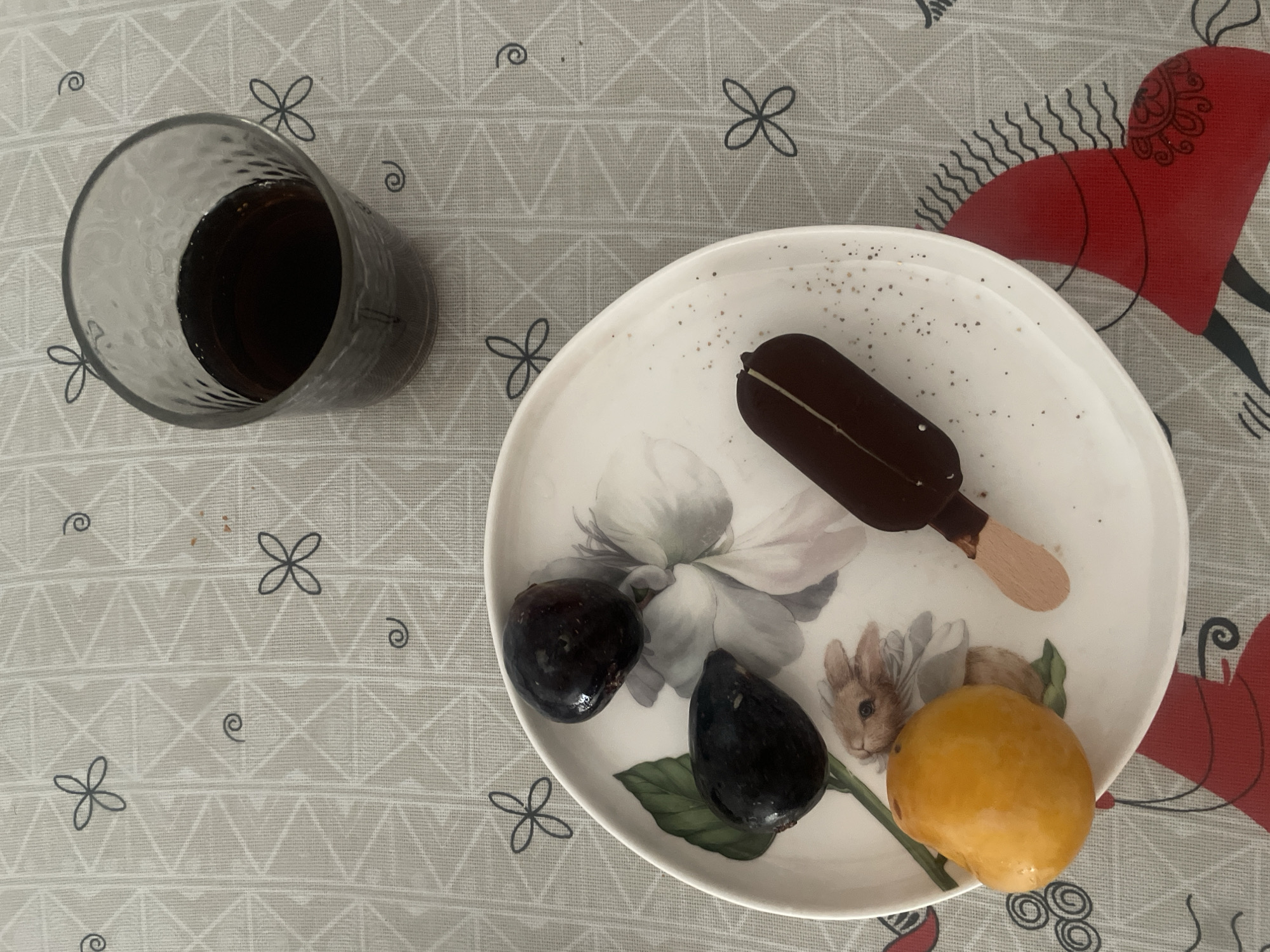 A close-up, overhead shot shows a white plate with a floral and rabbit design. On the plate are two dark purple figs, one orange fruit, and a chocolate-covered ice cream bar on a wooden stick. To the upper right of the plate, a glass filled with a dark liquid, likely cola, sits on a tablecloth. The tablecloth has a geometric pattern of interconnected triangles and is adorned with small dark gray flowers and swirls. A portion of a red and black illustrated horse is visible at the bottom right of the frame. The scene appears to be an indoor setting, possibly a dining table, and there is no discernible connection to the city of Atarfe, Spain, based on the visual information provided. There are no people or actions depicted.