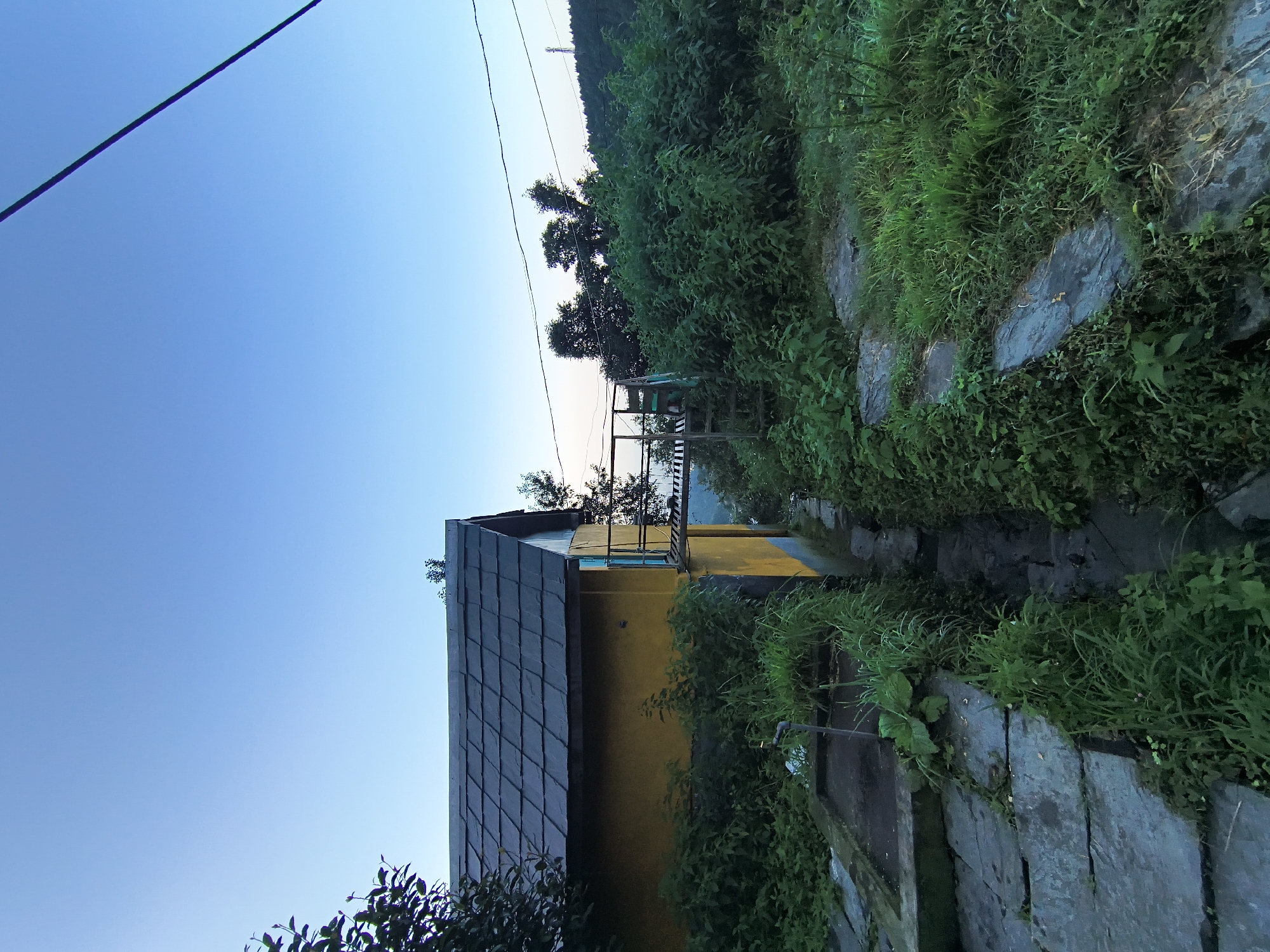 The image depicts a rustic scene on a hillside in Haini, India, under a clear blue sky transitioning to dusk. A yellow-painted building with a dark, tiled roof is partially visible on the left. In front of the building, a stone pathway leads past overgrown green vegetation. To the right of the building, a wooden staircase ascends towards a small balcony or landing area, surrounded by dense foliage. Further into the background, rolling hills covered in trees recede into the distance, with power lines crisscrossing the sky. The overall impression is one of a remote, natural setting with a simple dwelling.
