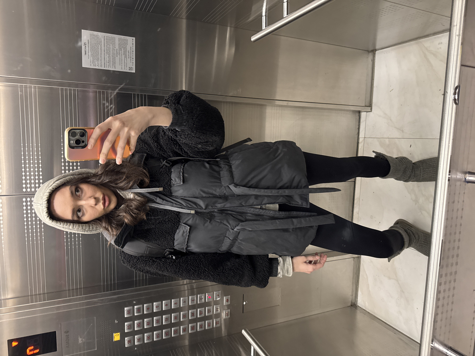A young woman takes a mirror selfie in an elevator. She is wearing a gray hooded beanie, a black fleece jacket, a black puffer vest, black leggings, and gray socks. The elevator has a stainless steel interior with a metal handrail. A button panel with numbers and Braille is visible. A sign is affixed to the wall above the woman. The button panel has labels in Russian. Based on the label AMDI2, the elevator is located in the city of Khimki, Russia.
