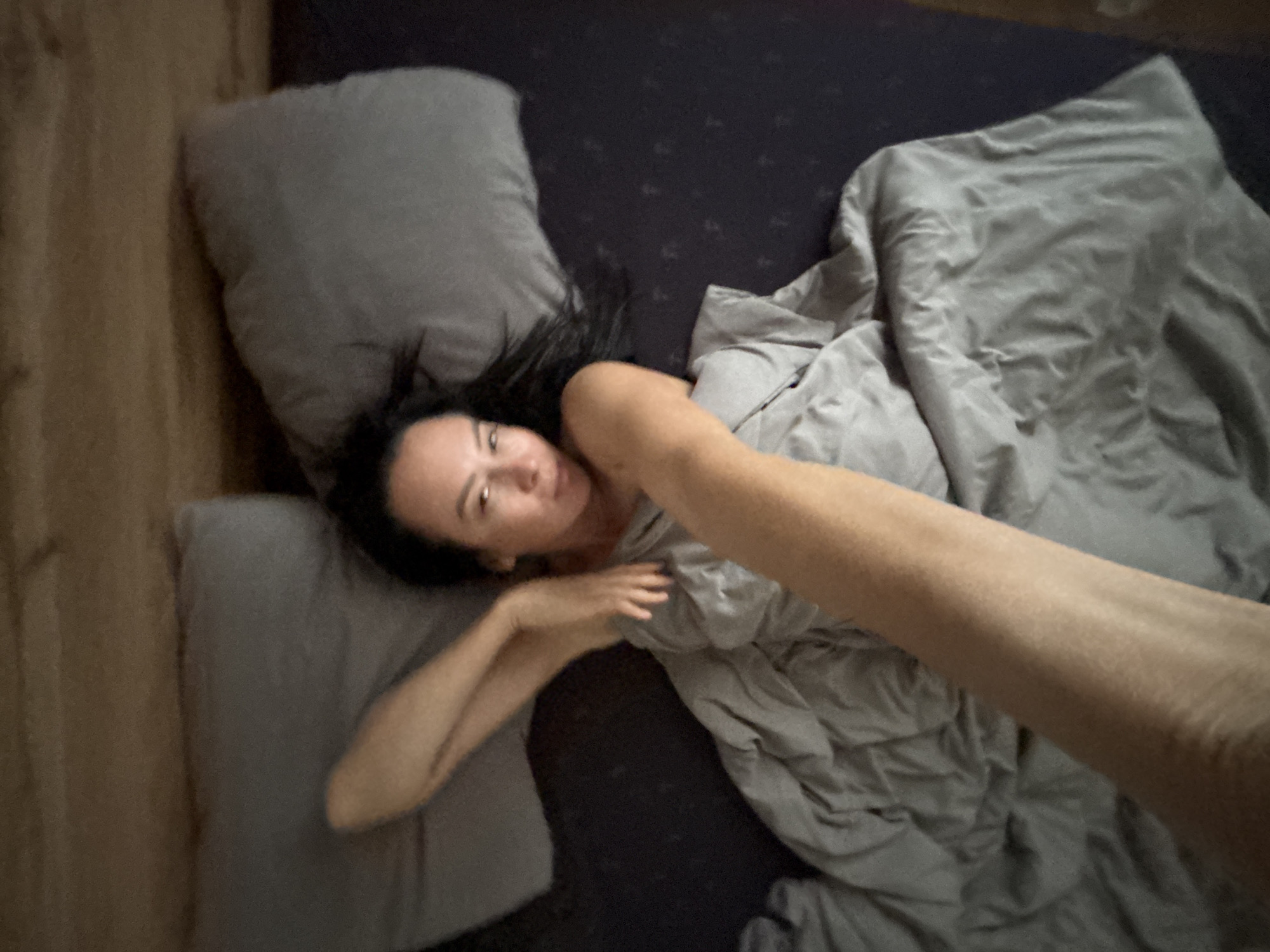The image shows a woman lying in bed, apparently taking a selfie. The bedding is gray, with two pillows at the head of the bed. The bed has a dark purple blanket on it. The woman has dark hair and is partially covered by a gray duvet. The photo was taken from an elevated angle, presumably by the woman herself. There is a wall with wood paneling visible on the left side of the frame. The location of Chaikovsky, Russia, cannot be independently verified from the given image.