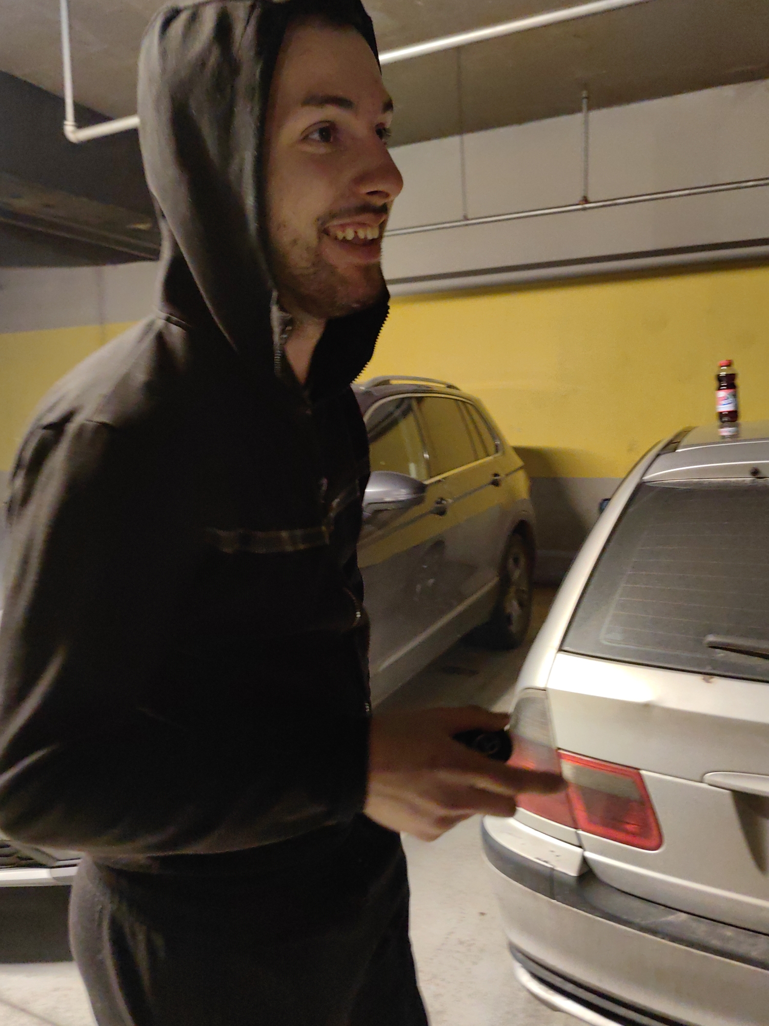 The image shows a man with dark hair and a stubble beard, wearing a black hooded sweatshirt. He is smiling and looking to the right. His right hand is partially visible and seems to be holding a dark object, possibly a car key. He appears to be standing in a parking garage, as suggested by the presence of two cars, one gray and one goldish-silver, and a yellow wall in the background. A bottle of liquid is standing on the roof of the silver car. The location cannot be determined to be in Sofia, Bulgaria.