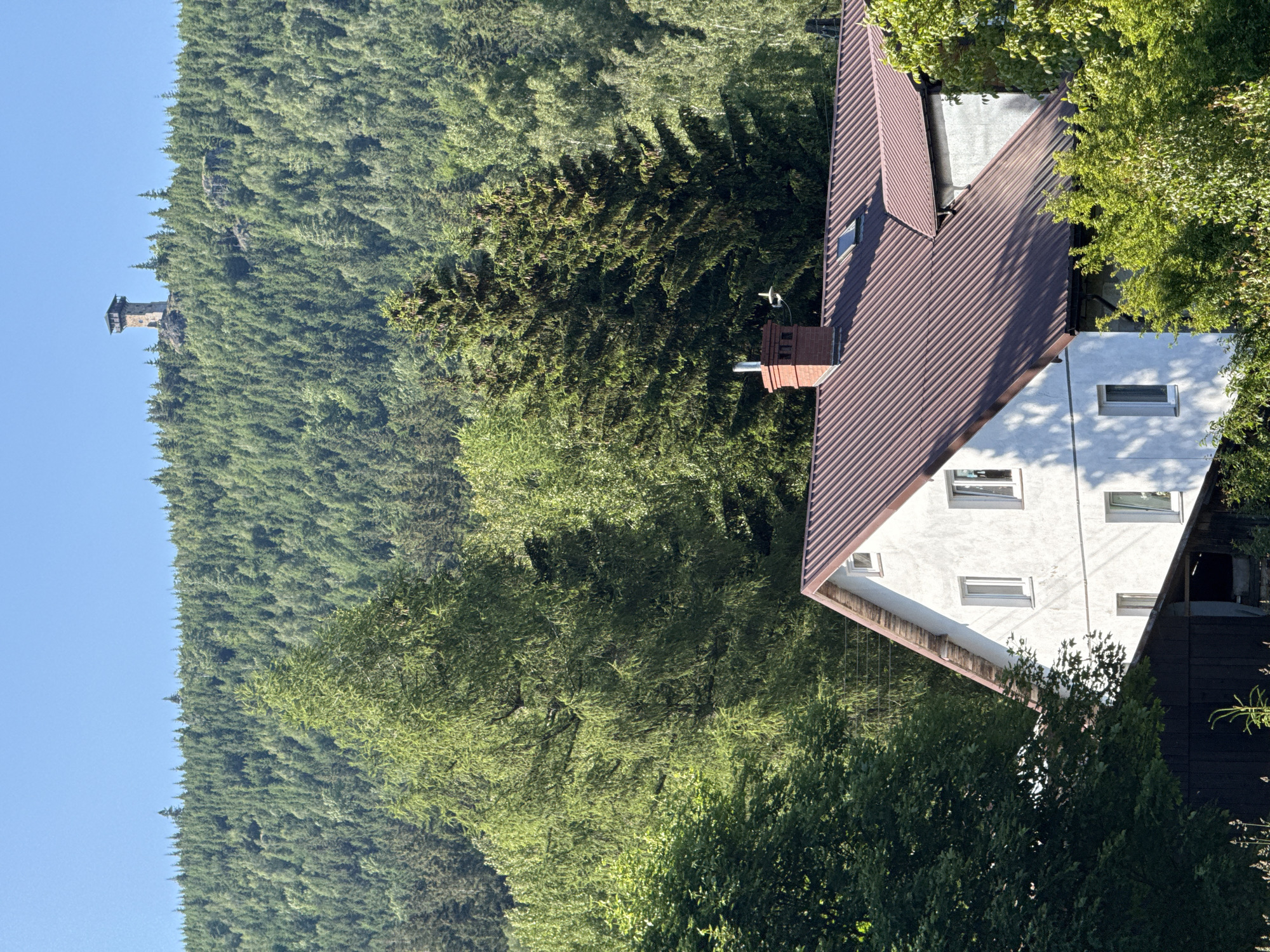 The image shows a building with a reddish-brown metal roof in the foreground, nestled amongst lush green trees. Behind the building, a dense forest covers a steep hillside, reaching up to the horizon. At the very top of the hill, visible in the distance, is a stone tower with a wooden observation deck. The sky above is a clear, bright blue. The scene appears to be a rural or mountainous area. The city of Szklarska Poręba is known for its natural beauty and is situated in a valley surrounded by the Karkonosze Mountains, which are heavily forested. The presence of the tower suggests it may be a lookout point or a historical structure within this natural landscape.