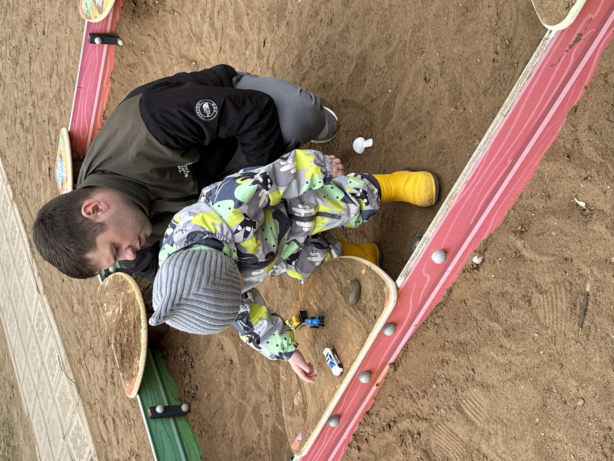 The image shows a man and a child playing in a sandbox. The man is wearing a black and green jacket and gray pants. The child is wearing a gray and green patterned snowsuit, a gray knit hat, and yellow boots. They are playing with toy cars in a wooden sandbox structure with red, green, and tan painted sections. The sandbox is filled with sand. The photo was taken in Chaikovsky, Russia.
