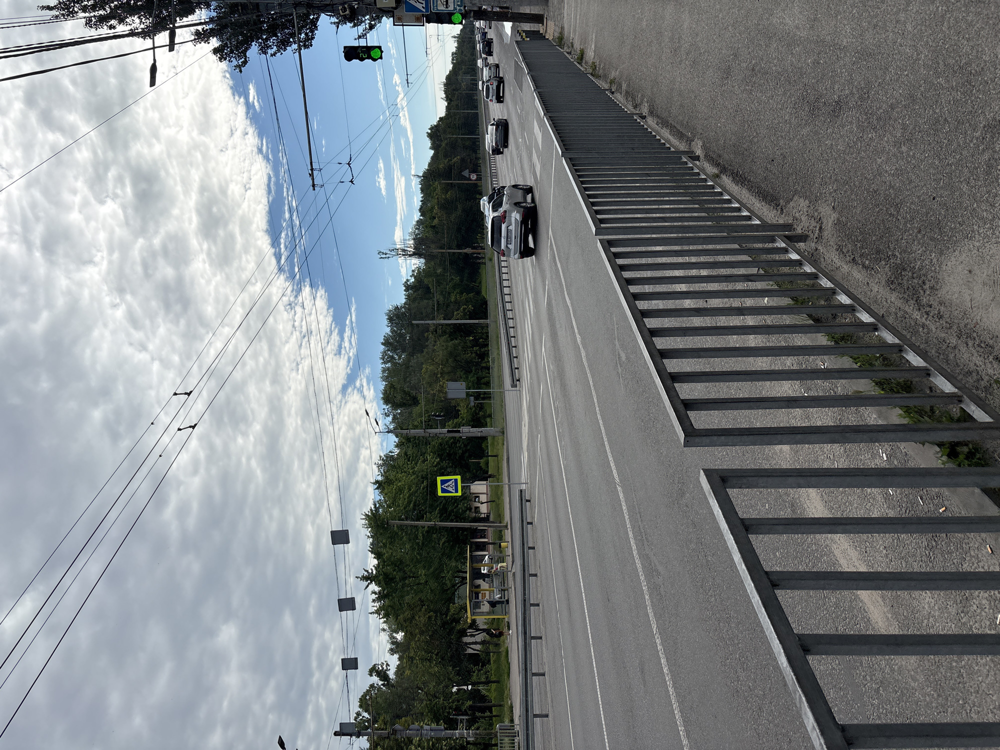 The image depicts a street scene in Dnipro, Ukraine. A multi-lane road runs horizontally across the frame, with a pedestrian walkway on the right, separated by a metal fence. Several cars are visible on the road, with some in motion and others stationary. A pedestrian crossing is marked on the road. On the left side of the road, a bus stop shelter with a yellow roof is visible, with a few people standing or sitting inside. Trees line the background, forming a dense green canopy. Overhead, numerous power lines crisscross the sky, which is partly cloudy. Traffic lights, some showing green, are mounted on poles along the road. A pedestrian crossing sign is also visible. The overall impression is of a typical urban street on a partly cloudy day.