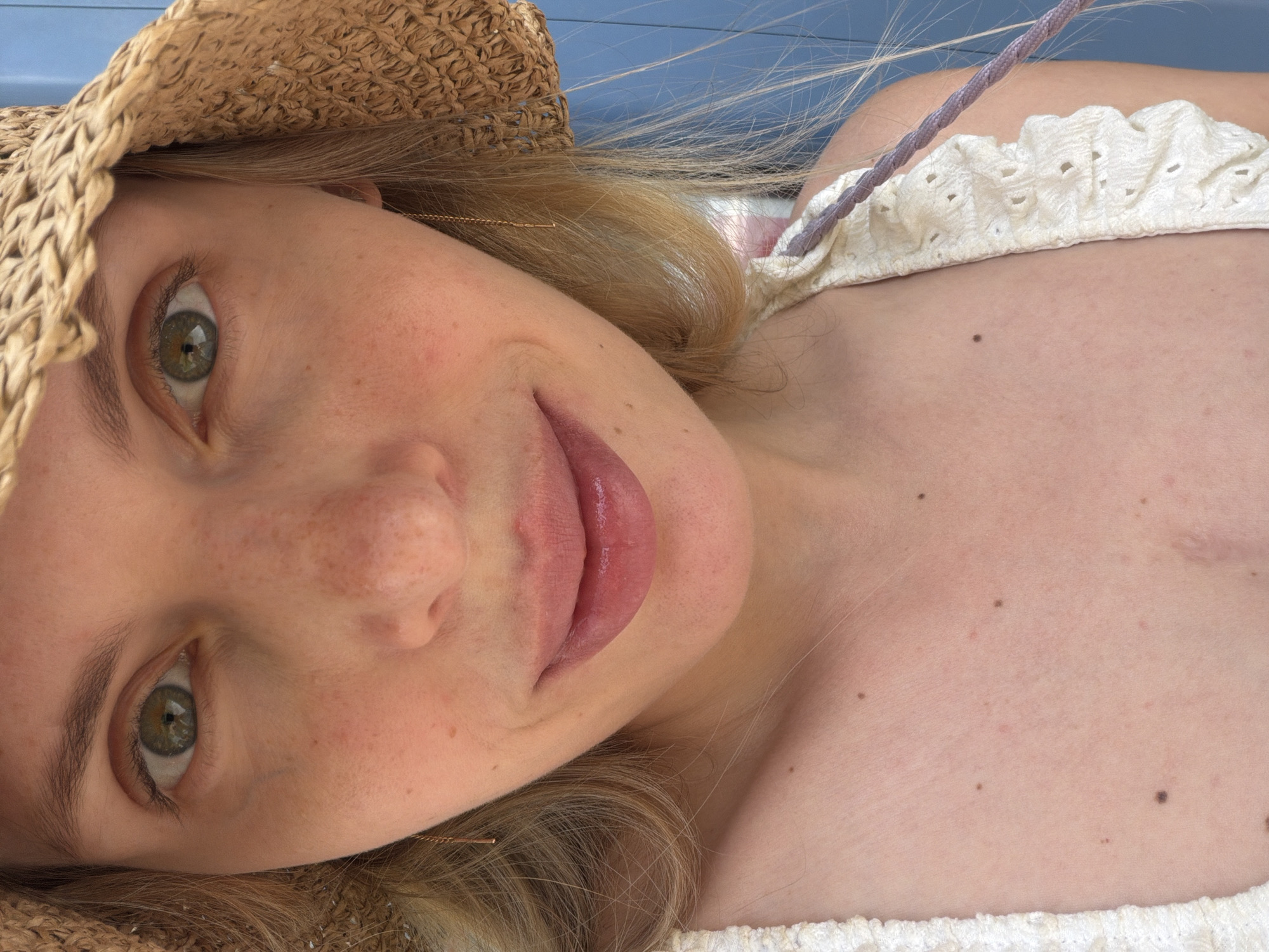 A close-up, high-angle shot shows a fair-skinned person with blonde hair and freckles wearing a woven straw hat and a white, lace-trimmed top. The person has green eyes and is smiling slightly. A faint scar is visible on the person's chest. No specific location in Budva, Montenegro is evident in the image.