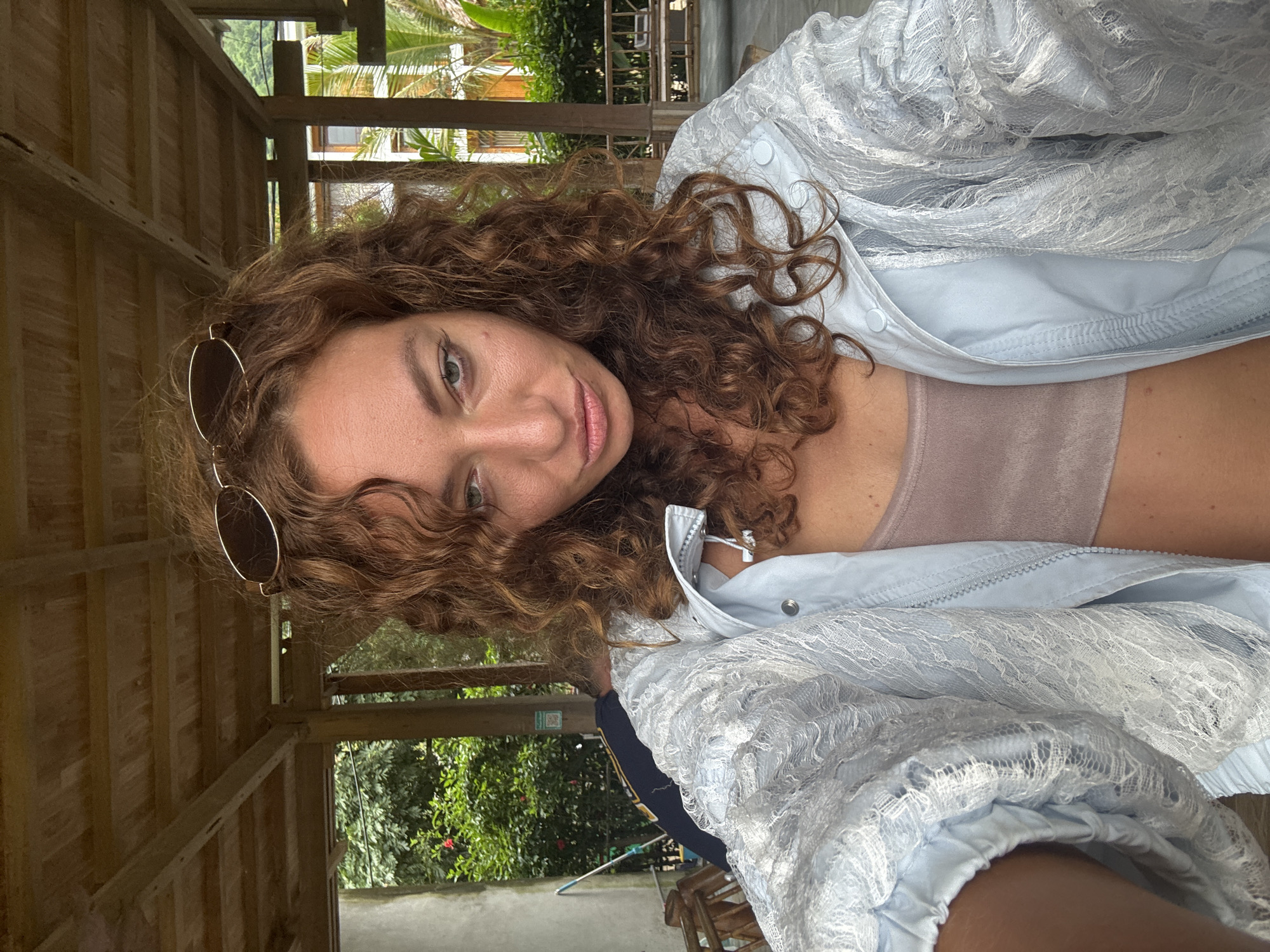 A woman with curly brown hair and sunglasses perched on her head is photographed from the chest up. She is wearing a light blue jacket over a taupe crop top. The background shows a wooden structure, lush green foliage, and a glimpse of a building. The scene appears to be outdoors, possibly in a tropical or subtropical location. The lighting suggests it is daytime.