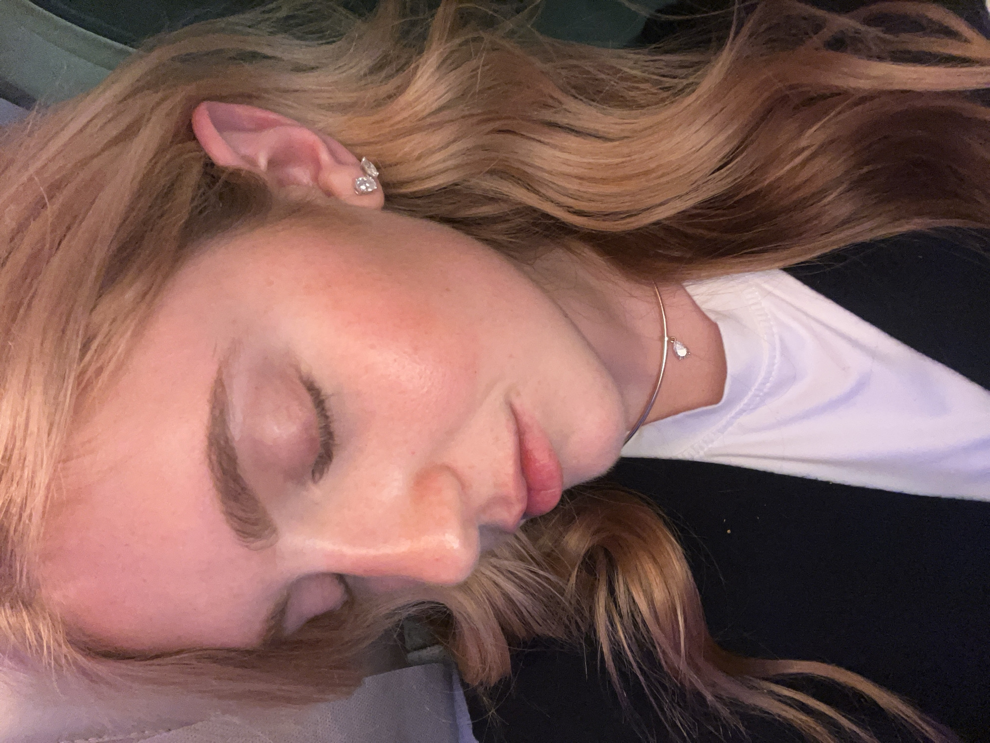 A close-up of a sleeping woman. Her face is pale with a pinkish hue on her cheeks. She has long, wavy, light brown hair with highlights. She is wearing earrings and a delicate necklace with a small charm. She is wearing a white t-shirt under a black jacket. The photo appears to have been taken indoors. No specific information suggests the location is in Kempton Park, South Africa. The location is unknown.