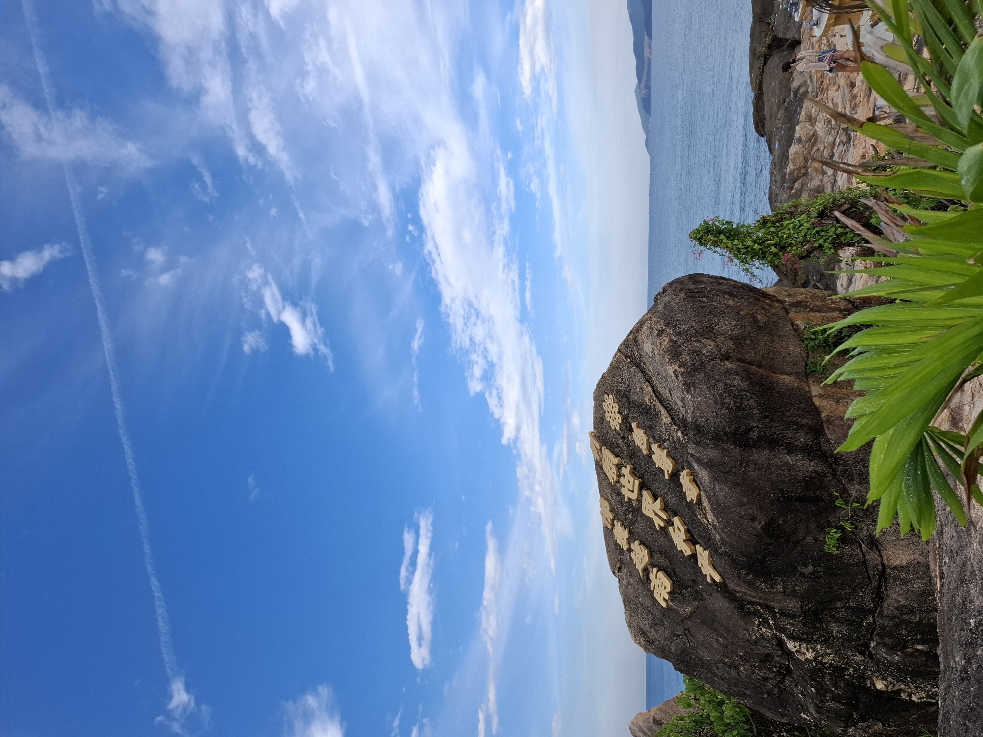 The image depicts a rocky coastal landscape under a bright blue sky with scattered clouds. A large, dark rock formation dominates the foreground, bearing raised, golden Chinese characters. The characters are arranged in two rows, with the top row containing four characters and the bottom row containing five, plus a decorative circular motif to the right of the bottom row. These characters are likely Buddhist sutras, commonly found at sites like Long Son Pagoda in Nha Trang, Vietnam, a popular tourist attraction known for its large reclining Buddha statue and the impressive white Buddha statue on a hilltop.
The sea stretches out in the background, meeting a distant, hazy coastline with mountainous terrain. In the middle ground, to the right of the large rock, there is a smaller rock outcrop covered in green vegetation, including vines with small pink flowers. A pathway or seating area is visible on the rocks to the far right, where a person, appearing to be a woman with dark hair tied back, is standing and looking towards the sea. She is wearing a light-colored top and shorts. In the immediate foreground, lush green palm leaves partially obscure the view. The overall impression is of a scenic, tranquil location with cultural significance.