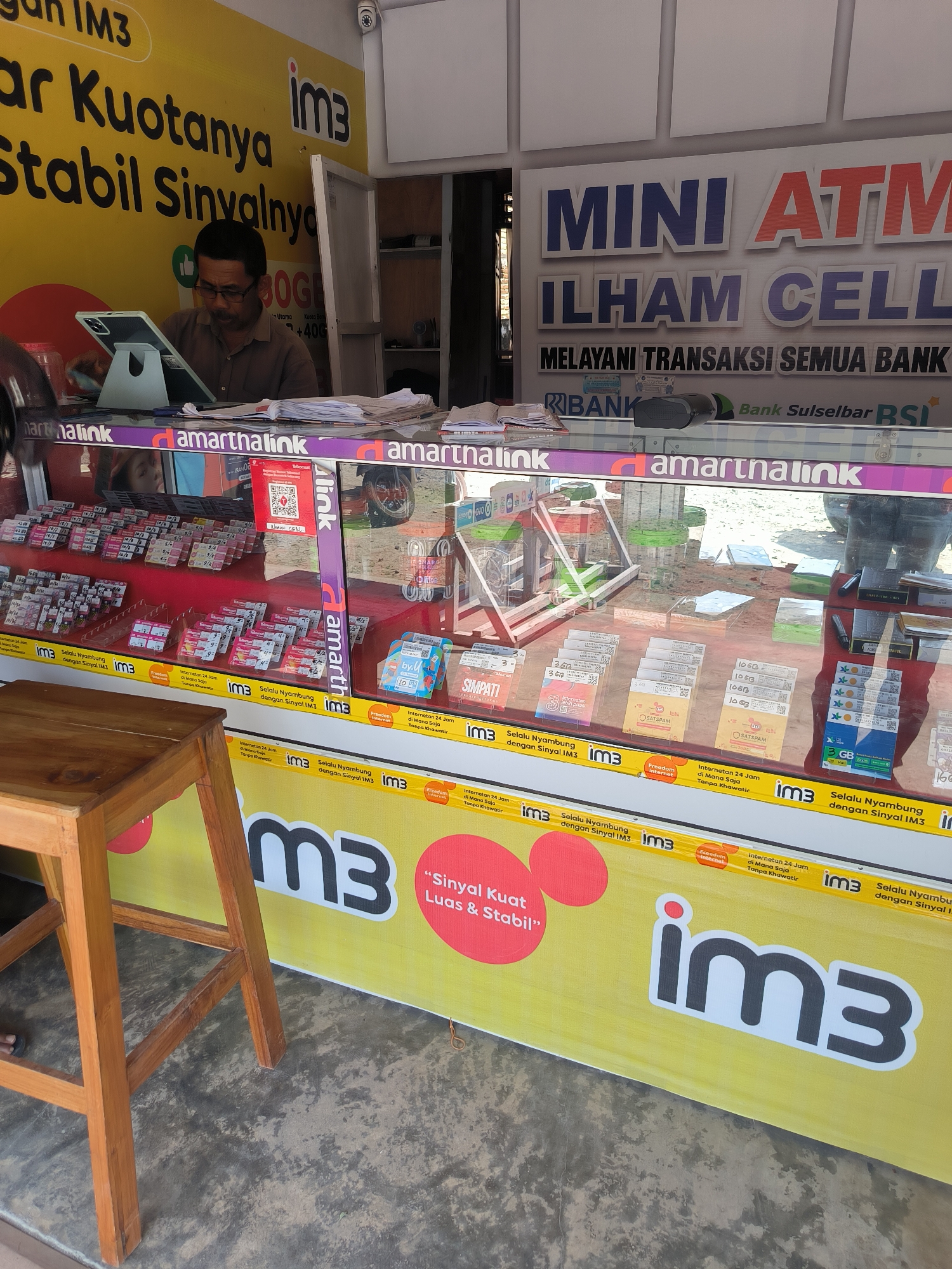 The image captures the exterior of a retail establishment in Lemo, Indonesia, likely a small business selling mobile phone services and related products. The storefront is predominantly yellow and adorned with prominent branding. Large white text on the yellow background reads "im3" and "Selalu Nyambung dengan Sinyal IM3" (Always Connected with IM3 Signal). A red graphic with white text states "Sinyal Kuat & Stabil" (Strong & Stable Signal).
A man wearing glasses and a brown shirt is seated behind a counter, looking at a tablet device. He appears to be an employee or proprietor. The counter is covered with various items, including stacks of papers and a tablet on a stand.
Behind the counter, a glass display case showcases a variety of mobile phone credit and data packages. These packages are neatly arranged in rows and appear to be from different mobile carriers, indicated by logos such as "im3" and "Simpati". Some packages are labeled with data amounts like "3 GB" and "10 GB".
Above the display case, a banner features large text that reads "MINI ATM ILHAM CELL MELAYANI TRANSAKSI SEMUA BANK" (MINI ATM ILHAM CELL SERVING ALL BANK TRANSACTIONS). This suggests the establishment also functions as a mini ATM service. Below this banner, logos for "BANK Sulselbar BSI" are visible.
The environment appears to be an outdoor setting, with a concrete floor visible in the foreground. A wooden stool is placed to the left of the frame, suggesting a seating area for customers. The overall impression is that of a busy local business serving the communication and financial needs of the community in Lemo.