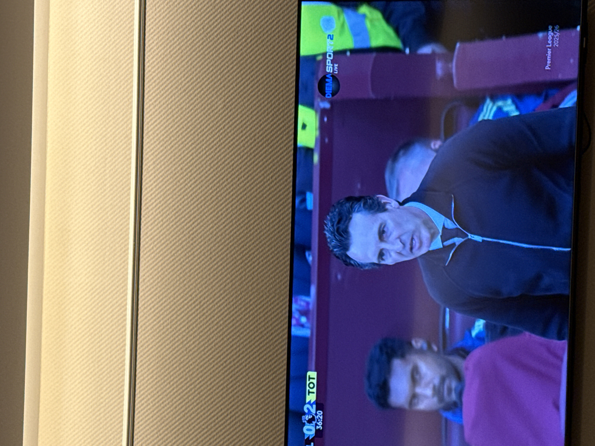 The image shows a television screen displaying a soccer game. The score is 1-2 in favor of Tottenham, and the time is 36:20. The broadcast is on Diema Sport 2 Live. The Premier League logo and the year 2025/26 are visible in the top right corner of the screen. There are three men on the screen, two with darker complexions and one lighter skinned. The man in focus appears to be a soccer coach. The image appears to have been taken indoors in a home.