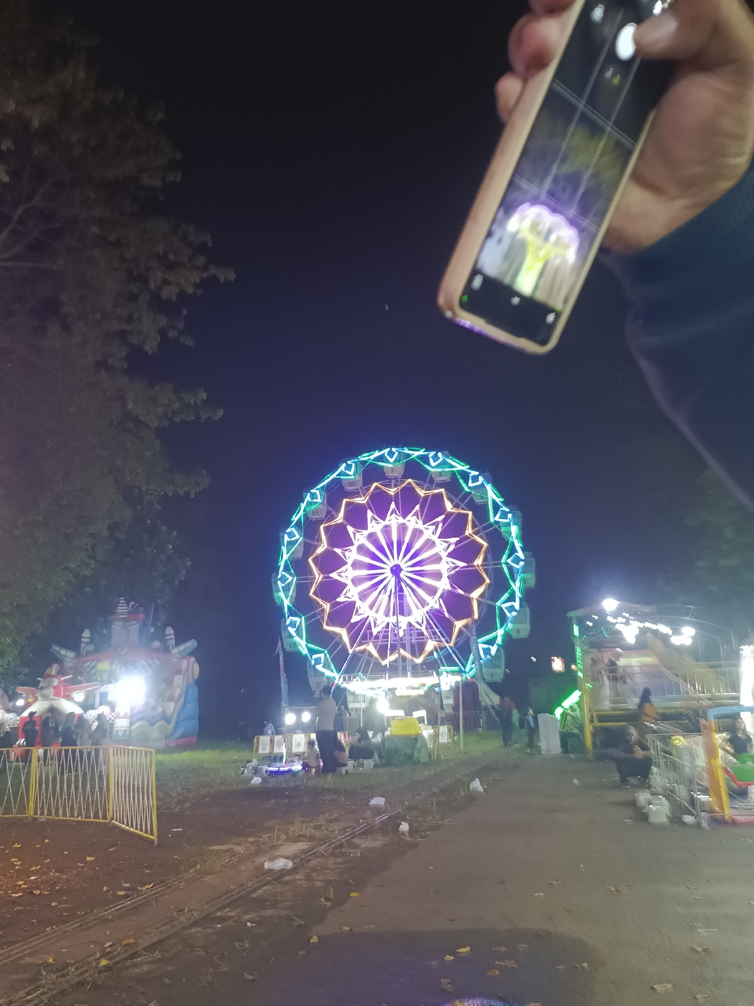 The image captures a nighttime scene in Tulangan, Indonesia, featuring a brightly lit Ferris wheel as the central focal point. The Ferris wheel is decorated with purple, white and green lights. Several other fairground attractions and structures are visible in the background, illuminated by bright lights. A dirt path is visible in the foreground, and a tree is visible to the left of the Ferris wheel. A person is holding up a smartphone in the upper right corner, displaying an image of a tree illuminated by green lights on its screen. The sky is dark, indicating it is nighttime. A metal fence is visible in the lower-left corner. The ground near the path is covered with fallen leaves. People are seen near the Ferris wheel and the other attractions.
