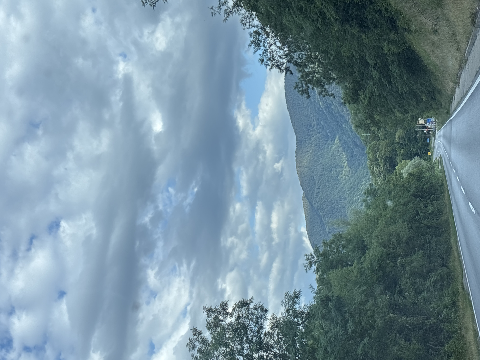 The image depicts a scenic road leading towards a mountainous landscape under a cloudy sky. The road is paved and appears to be a two-lane highway with white dividing lines. Lush green trees line both sides of the road, with dense foliage creating a tunnel effect. In the distance, rolling hills covered in trees rise up to meet the sky, which is filled with dramatic, textured clouds. There are no people visible in the image. The location is identified as the city of Zlatirat, Bulgaria. The scene suggests a rural or mountainous area, likely a popular route for scenic drives. The weather appears overcast, but not actively raining.