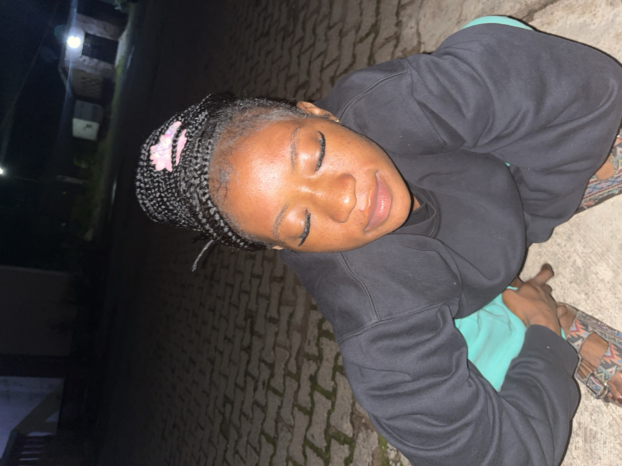 A dark-skinned person with braided hair, wearing a black sweatshirt and teal pants, is crouching on a paved ground, possibly a street or walkway, at night. The person's eyes are closed, and they appear to be looking down. The background is dimly lit, with hints of buildings and streetlights, suggesting an urban or suburban environment in Jos, Nigeria. The person is wearing patterned sandals. The lighting is low, and the image is captured from a slightly elevated angle.