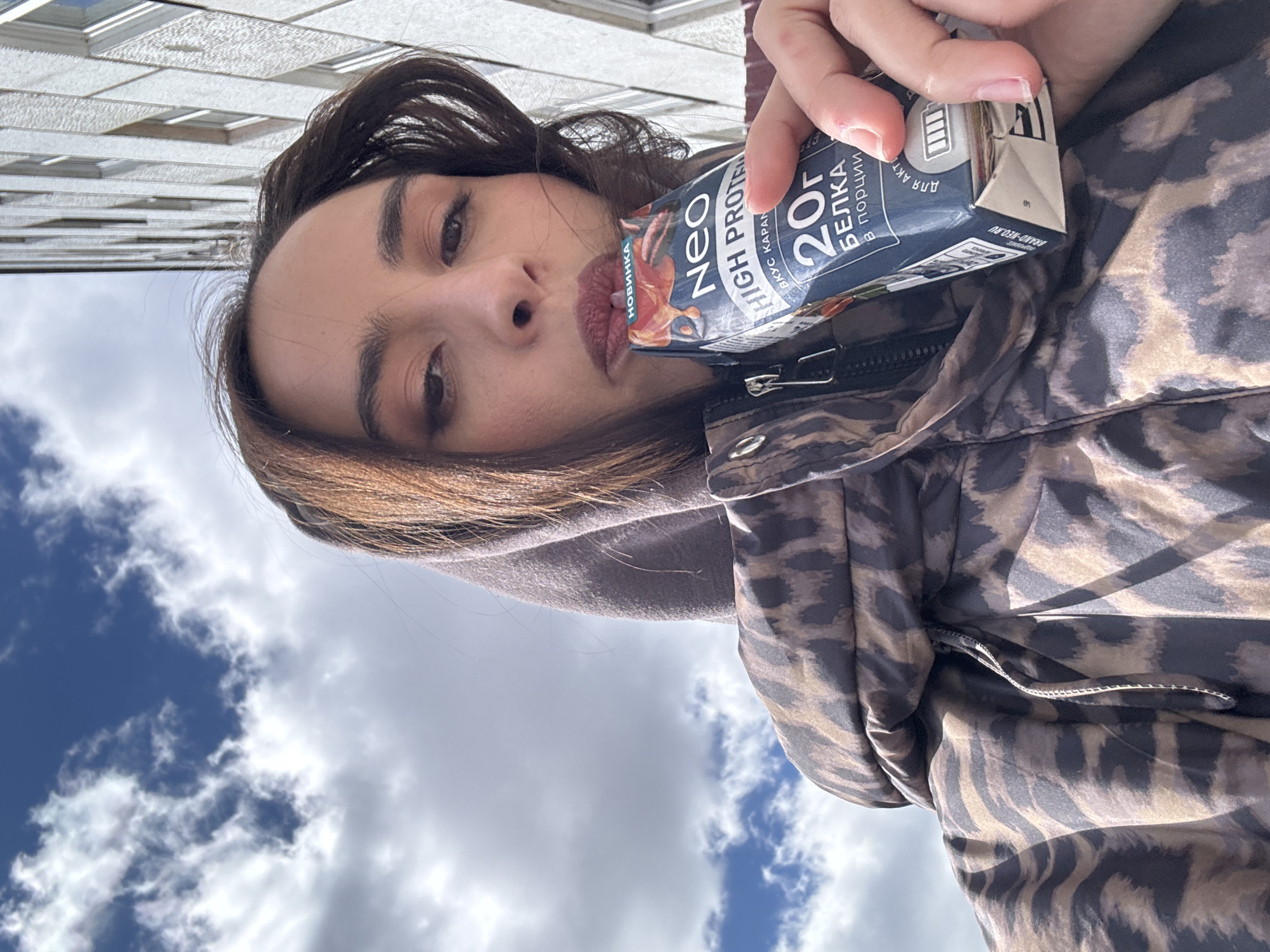 A woman with dark hair and brown eyes holds a "Neo High Protein" drink up to her lips. The drink's packaging is blue and white, with text in Russian. The woman is wearing a brown and tan animal print jacket. Behind her is a building and a sky with white clouds. The image appears to be taken from a low angle, looking up. Without additional information, determining if the image was taken in Krasnoyarsk, Russia, is impossible.