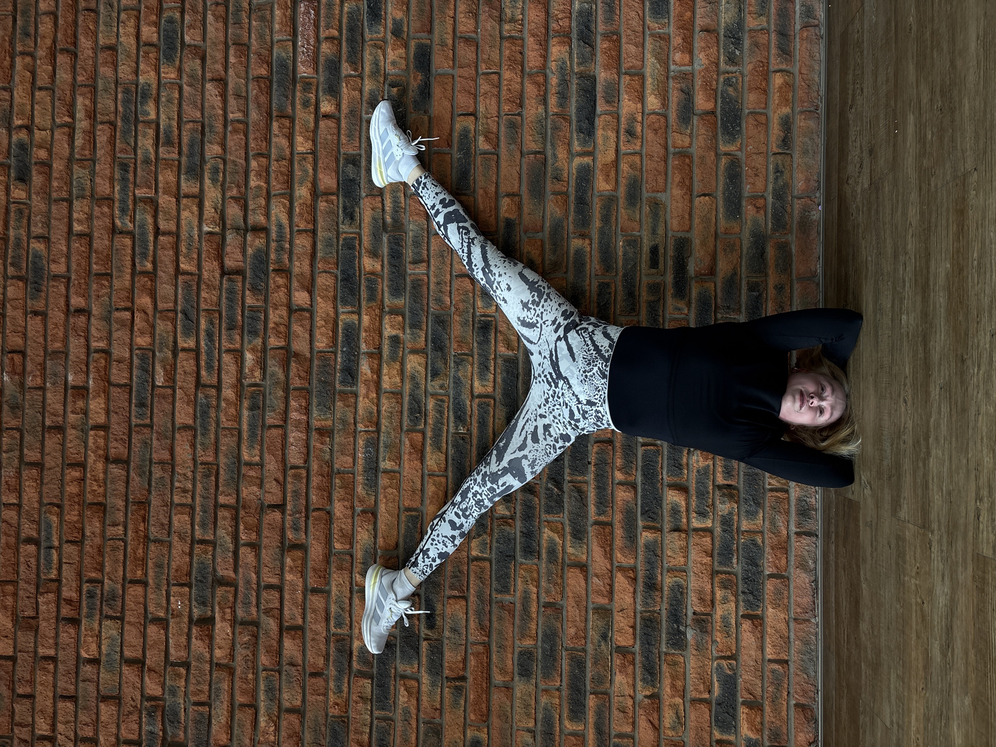 A woman lies on her back on a brick floor, with her legs spread in a V-shape. She wears a black long-sleeved shirt and patterned leggings, along with gray sneakers. Her arms are positioned behind her head. The brick floor is a mix of red and brown tones. To the right of the brick floor, there is a wooden surface. There is no evidence to suggest the location is the city of Chaikovsky, Russia.