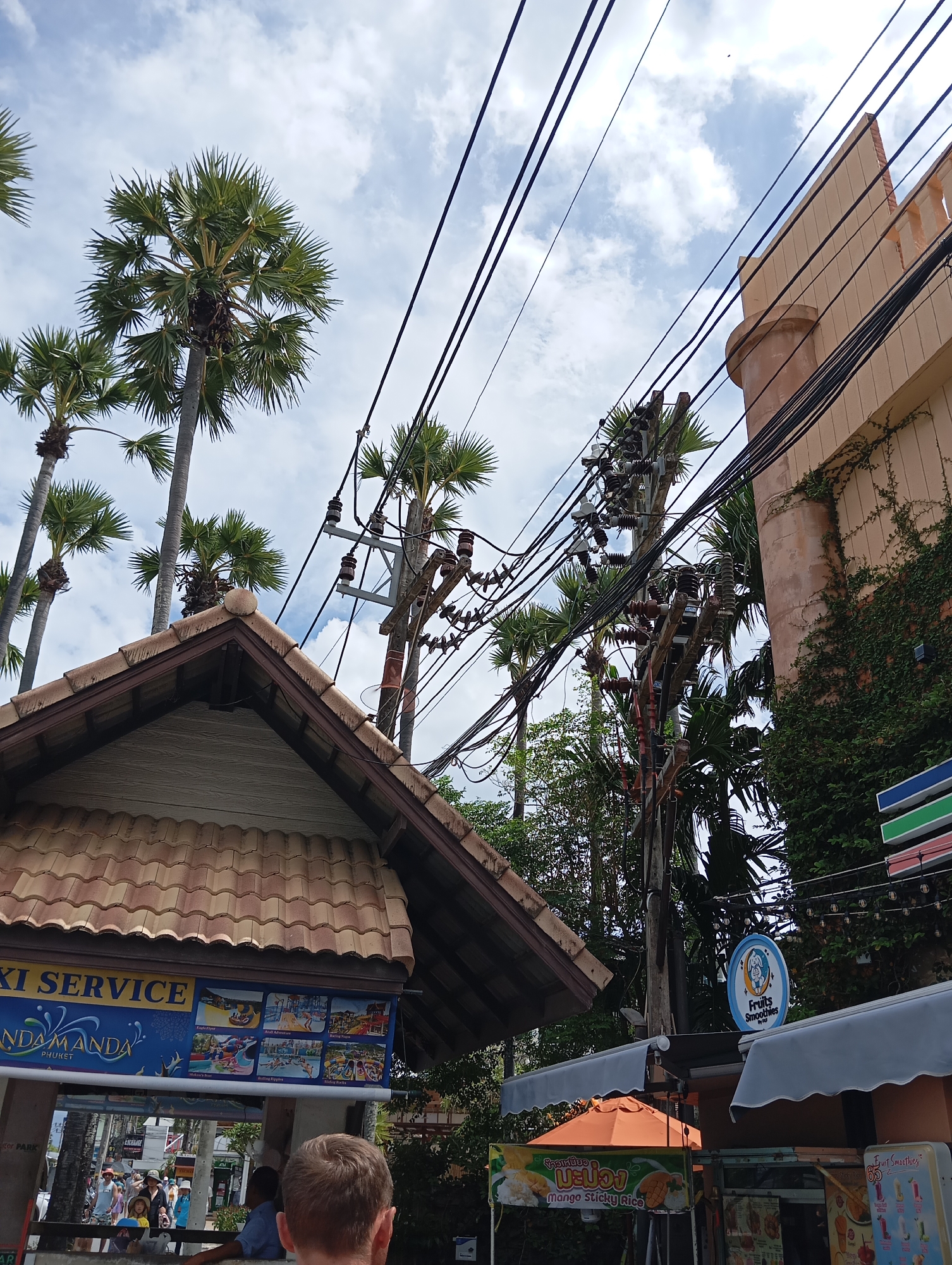 A street scene in an urban environment, likely a tropical location, is captured from a low angle looking upwards. The sky is partly cloudy with a bright white and blue hue. Dominating the frame are numerous power lines and utility poles, with a complex network of black wires stretching across the sky.
In the foreground, the tiled roof of a building is visible, with a light-colored wooden siding underneath. To the left of the roof, a sign advertises "TAXI SERVICE" and "ANDAMANANDA PHUKET" with images of water park attractions. Further down the street, several people are walking.
To the right of the frame, a light-colored building with an arched balcony railing and a cylindrical turret stands. Green ivy is growing up the side of this building. Below the building and to the left, a round blue sign reads "Fruits Smoothies."
In the midground, several tall palm trees are scattered throughout the scene, their fronds reaching towards the sky. Among these, a more detailed view of utility poles with insulators is visible, supporting the network of wires. Another sign, featuring an orange awning, displays "Mango Sticky Rice" in Thai and English.
The overall impression is a busy street in a tropical city, characterized by a dense urban infrastructure of power lines and palm trees, with signs indicating commercial establishments.