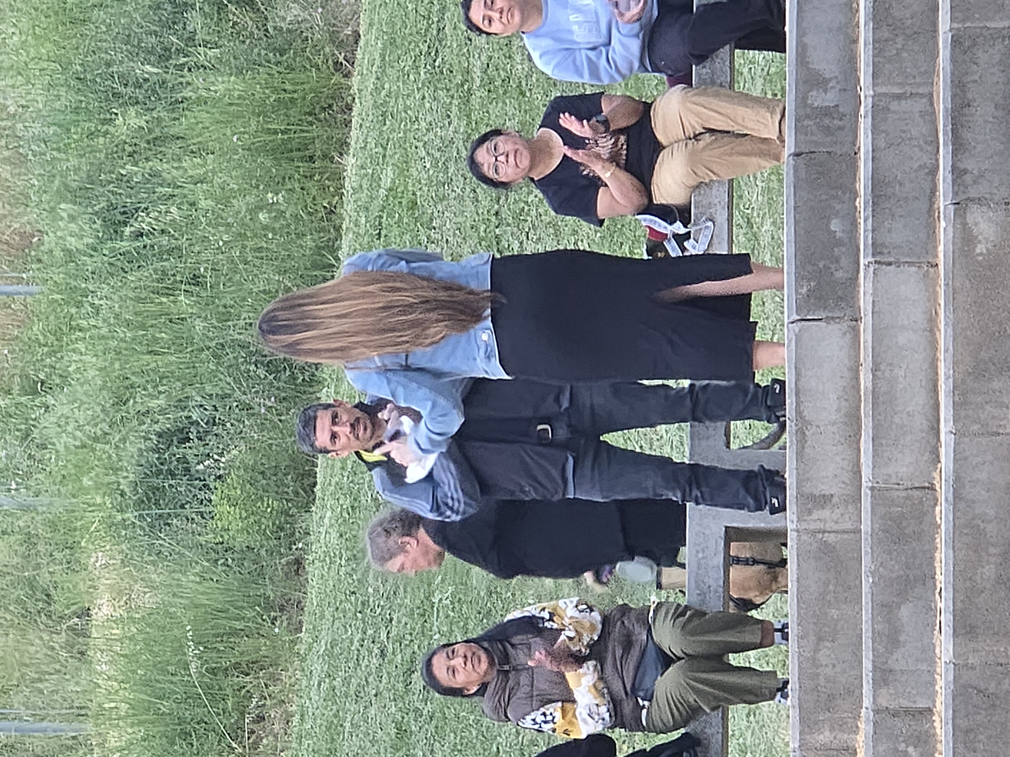 The image captures an outdoor gathering of at least nine individuals in Cornellà De Llobregat, Spain, during daylight hours. The setting features a prominent expanse of green grass on the left, contrasting with concrete steps or bleachers on the right, suggesting a public park, sports area, or amphitheater-like space.
In the foreground, a woman with long, light brown hair, dressed in a denim jacket and a black long skirt, stands with her back partially turned to the camera. To her right, a man in a light blue top and dark trousers stands, looking towards the right side of the frame. Below these two, another man in a dark jacket is visible. Further down and to the left, a woman in a distinctive patterned jacket (yellow, black, and white) and green cargo pants sits on the grass, also observing something to the right.
Behind the standing woman, an older woman with glasses, wearing a black t-shirt and tan pants, is seated on the concrete steps, actively clapping her hands. Above her, two other people are seated: one in a light blue sweatshirt and dark pants, and another person whose upper body is partially visible in a dark top. Another man in a grey sweatshirt and dark pants is also standing further up the steps.
The people are dressed in casual attire, indicating a relaxed atmosphere. The bright and clear lighting suggests it is daytime, possibly late afternoon. The group appears engaged, with the clapping woman indicating appreciation or participation in an ongoing activity or event. No discernible text is visible on clothing or in the background.