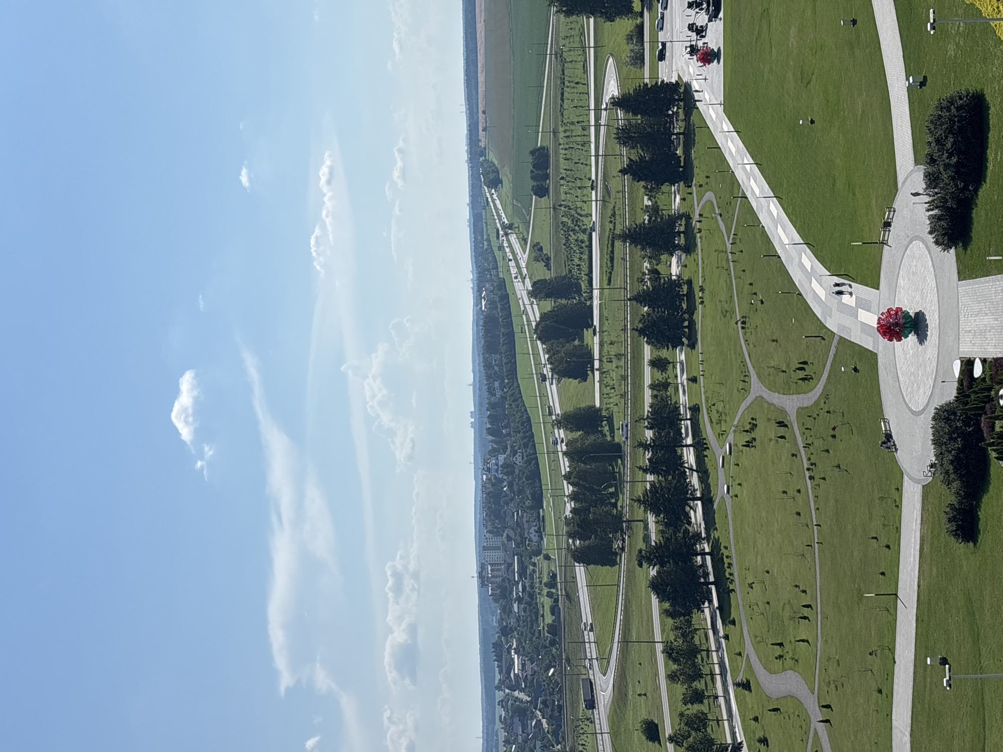 A high-angle, full shot captures a clear, bright day in a park-like area within the Ozyaritska-Slabadski rural council, Belarus. The foreground features a well-maintained green lawn with winding paved pathways. In the lower center, a large, spherical red sculpture is visible on a circular paved area. Further along a pathway, two people are walking. Another red sculpture is visible to the right, near a parking area with several cars.
The middle ground is characterized by more green spaces, scattered trees, and curving roadways that appear to be part of a highway interchange. Numerous cars are visible on the roads. The background reveals a sprawling urban or suburban landscape with various buildings, including a tall multi-story structure and several smaller houses, interspersed with more greenery and fields. The horizon is marked by a line of trees and distant structures. The sky is a vibrant blue with scattered white clouds.