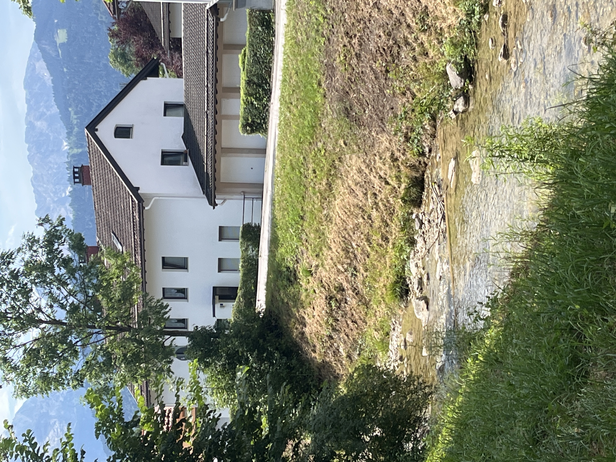 This image depicts a residential area in Garmisch-Partenkirchen, Germany, featuring a white house with a brown tiled roof and a driveway with garage doors. The house is situated on a slight incline, with a neatly trimmed hedge lining the property. A narrow, shallow stream with a rocky bed flows through the foreground, bordered by lush green grass. In the background, a lush green tree stands tall, and behind it, the majestic blue peaks of the Bavarian Alps rise under a clear sky. The scene appears serene and tranquil, suggesting a peaceful day in a picturesque mountain town.