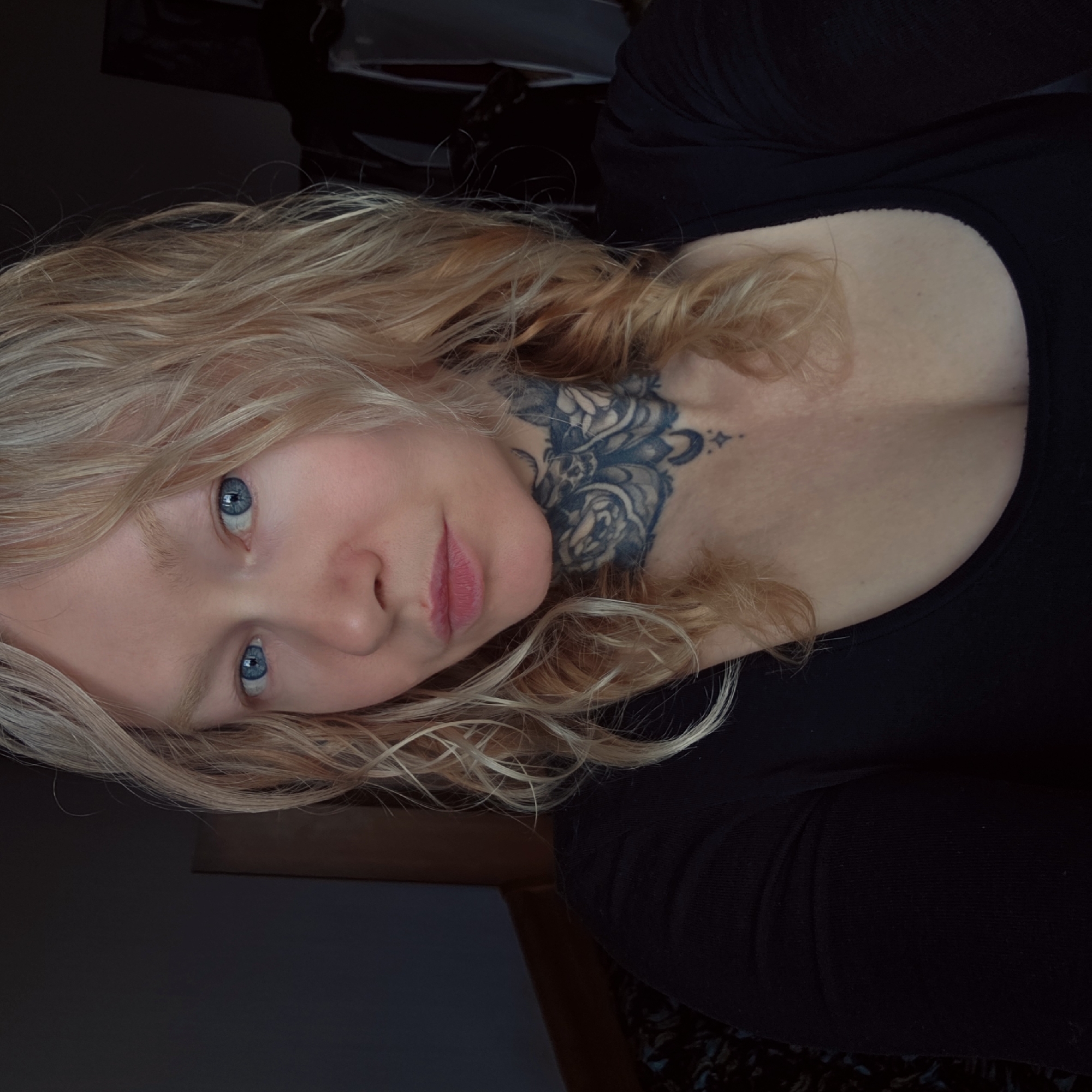 A close-up, angled portrait shows a fair-skinned person with blonde, wavy hair and striking blue eyes. They have a prominent nose and pink lips. A tattoo is visible on their neck, featuring a dark floral design with a crescent moon and a small star. The person is wearing a black top. The background is dimly lit and partially obscured, hinting at an indoor setting. There is no information about the city.