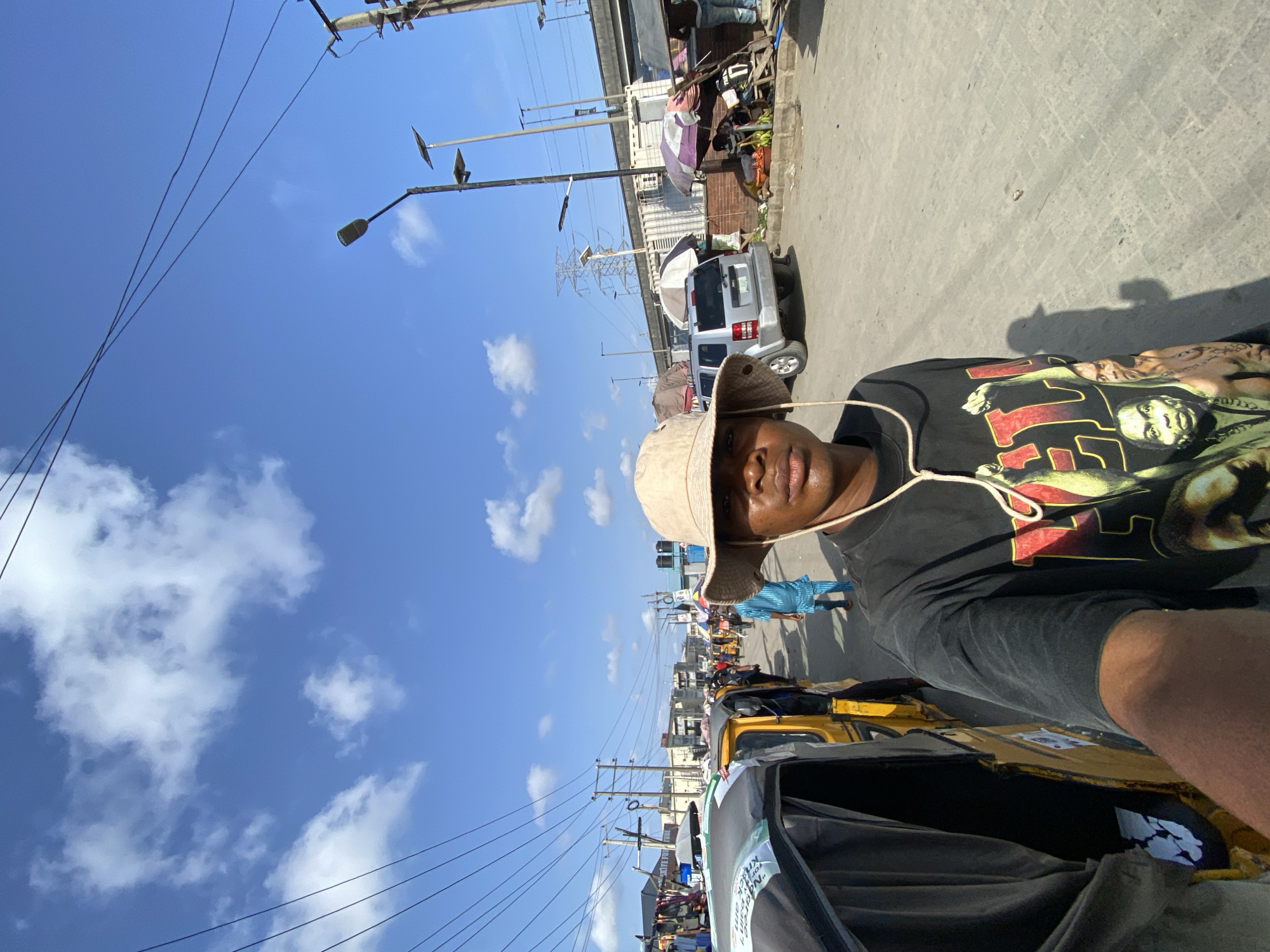 The image, taken in Lekki, Nigeria, shows an individual with dark skin, wearing a bucket hat and a black t-shirt with a graphic print. The person is likely riding in a "keke," a type of auto-rickshaw. The background shows a busy street with various vehicles, including a silver SUV, and pedestrians. Streetlights and power lines are visible against a blue sky with scattered clouds. Buildings and makeshift stalls line the side of the road.