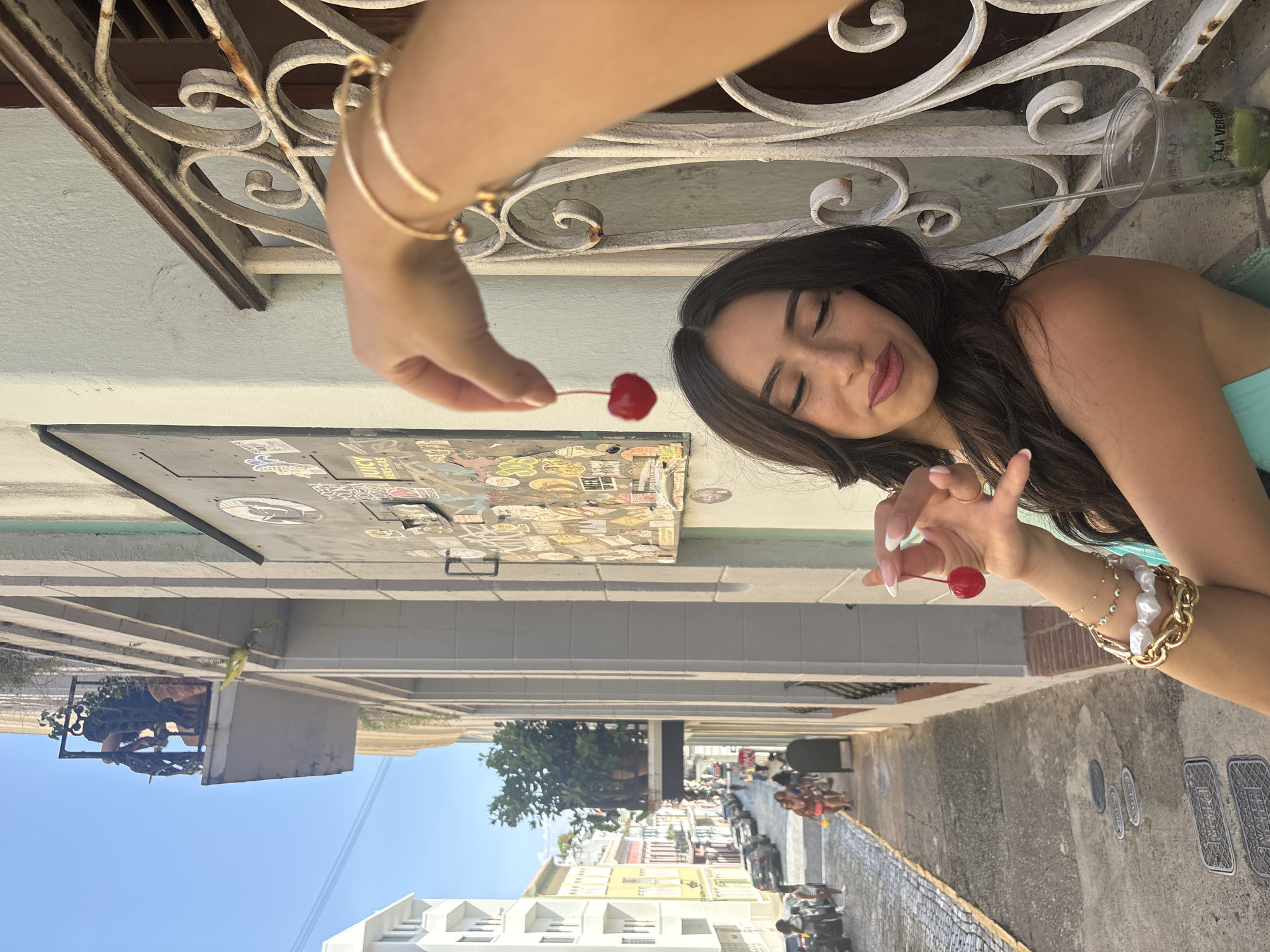 In San Juan, Puerto Rico, a young woman with dark, wavy hair is shown from the chest up, with her eyes closed and a slight smile. She is holding two cherries, one in each hand, in front of her face. She is wearing a light blue top and several bracelets on her left arm, including a chunky gold chain and pearl bracelet. To the right of the woman is a clear plastic cup with a green liquid and a straw, resting on a window ledge. In the background, a cobblestone street is visible with cars parked along the side and people walking in the distance. The architecture suggests an older part of the city, with buildings of various colors and balconies. The overall lighting indicates it is daytime with bright sunlight.