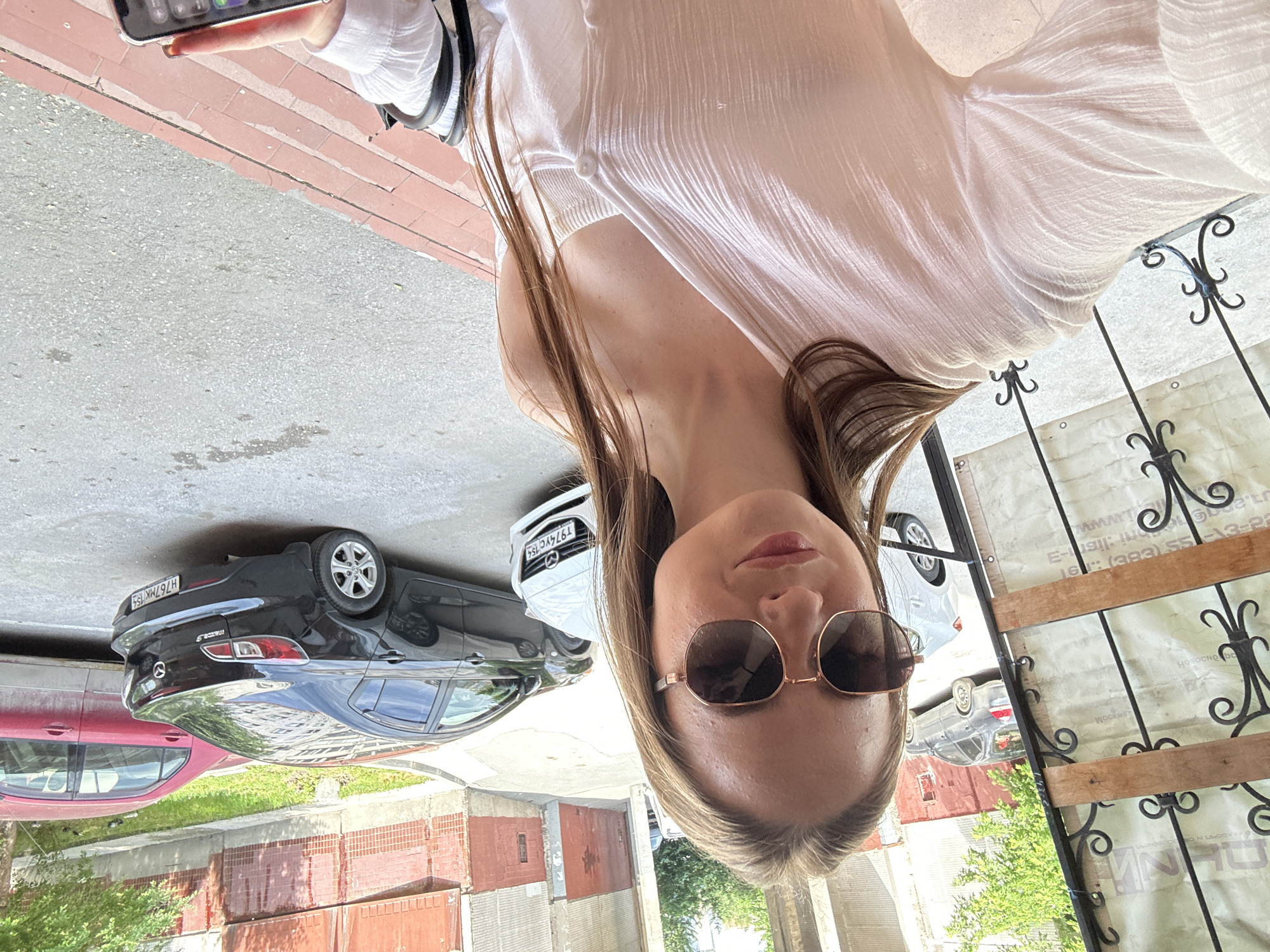 A young woman with long brown hair, wearing sunglasses and a white top, stands on a brick pathway in front of a building. She is holding a smartphone in her right hand. Behind her, several cars are parked on a paved street. The scene appears to be outdoors on a sunny day.