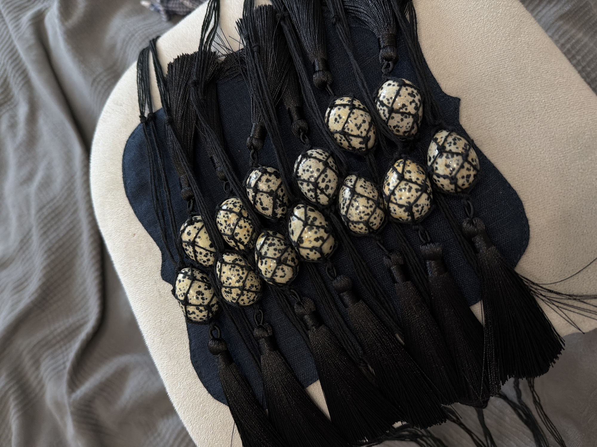 This media file displays a close-up, overhead view of several black tassels with decorative, egg-shaped beads. The beads have a cream-colored base with a speckled pattern of black dots and are intricately wrapped with black string, creating a geometric design. The tassels themselves are uniformly black and appear to be made of fine threads.
The objects are arranged on a surface that features layers of fabric. A dark navy blue fabric provides a base for the tassels and beads, and this is bordered by a lighter, off-white fabric with a textured surface. To the left of these fabrics, a gray, softly draped material is visible, suggesting a soft textile like a blanket or throw. The lighting appears to be neutral and diffused, without strong shadows, indicating it might be taken indoors or on an overcast day.
The arrangement of the tassels and beads suggests they might be decorative elements, possibly from a craft project, home decor item, or jewelry. There are no people or animals visible, and the focus is solely on the texture and pattern of the objects and their immediate surroundings. No text is visible in the image. The overall impression is one of detailed craftsmanship and a focus on tactile elements.