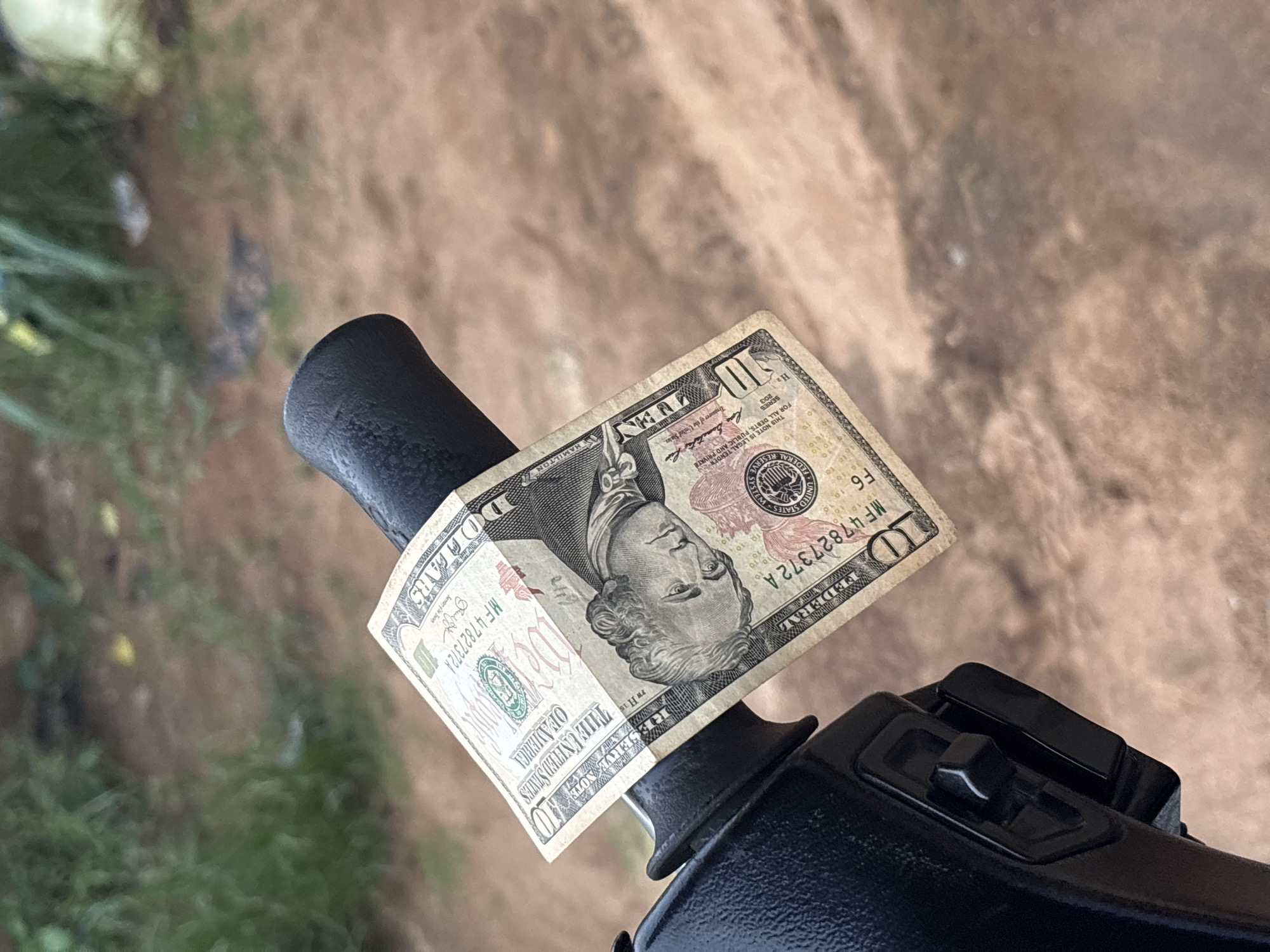 A ten-dollar bill is folded and placed over a black apparatus, possibly a lever or part of a machine. The bill shows a portrait of Alexander Hamilton and has the serial number MF47827372A. The background appears to be a blurred mix of outdoor elements, including possible vegetation and ground textures. The location, Ikot Ekpene, Nigeria, is not visually verifiable from the image content.