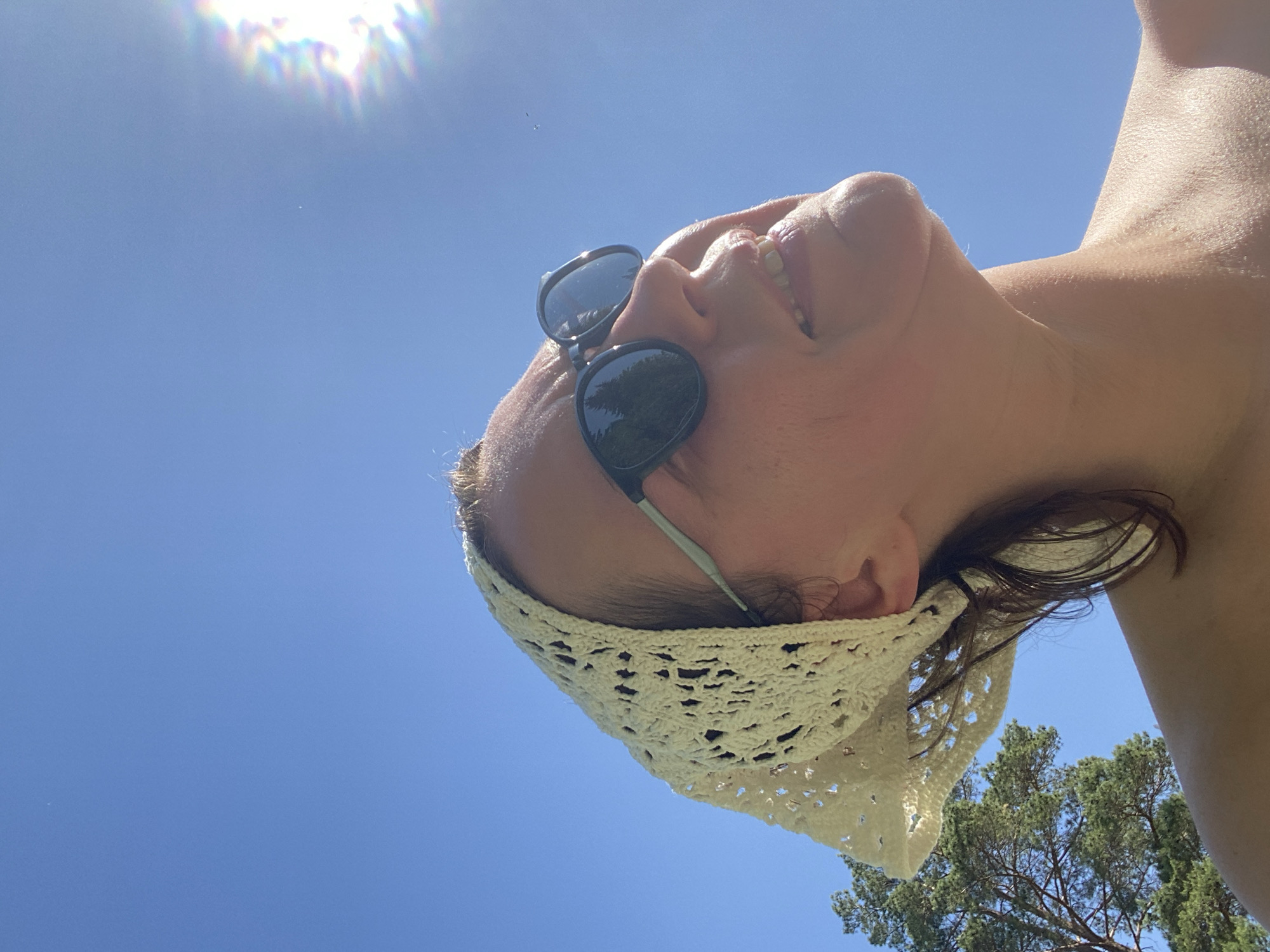 A low-angle shot captures a person, likely female, wearing sunglasses and a white, possibly crocheted, headband. The person is smiling, revealing their upper teeth. The background is a clear blue sky with a bright, out-of-focus light source resembling the sun. A portion of a tree is visible in the lower right corner. The location is not possible to determine based on the visual elements of this image.