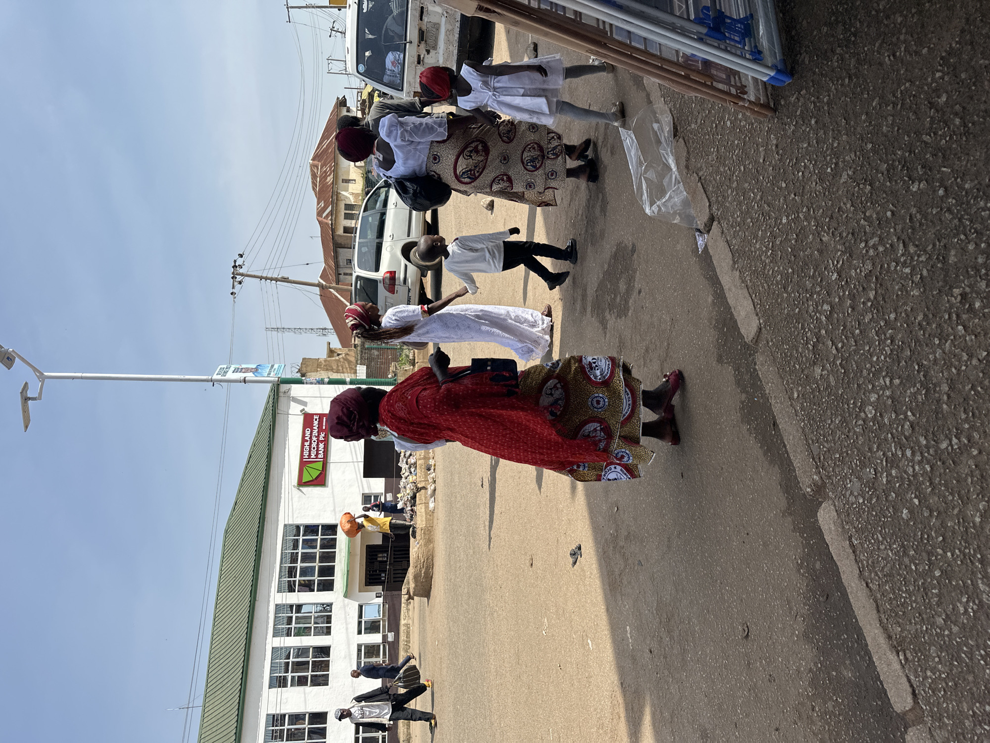 In Bukuru, Nigeria, on a sunny day, people walk along a paved street in front of a white building with a green roof, identified as Highland Microfinance Bank Plc. A group of individuals, including women in traditional attire and children, are walking on the street. One woman in the foreground is wearing a red dress and a headscarf, while another woman next to her wears a white dress with a patterned skirt. A young boy in a white long-sleeved shirt and dark pants walks beside them. In the background, other people are seen walking, and vehicles are parked on the street. The environment appears to be an urban setting with a mix of commercial and residential buildings.