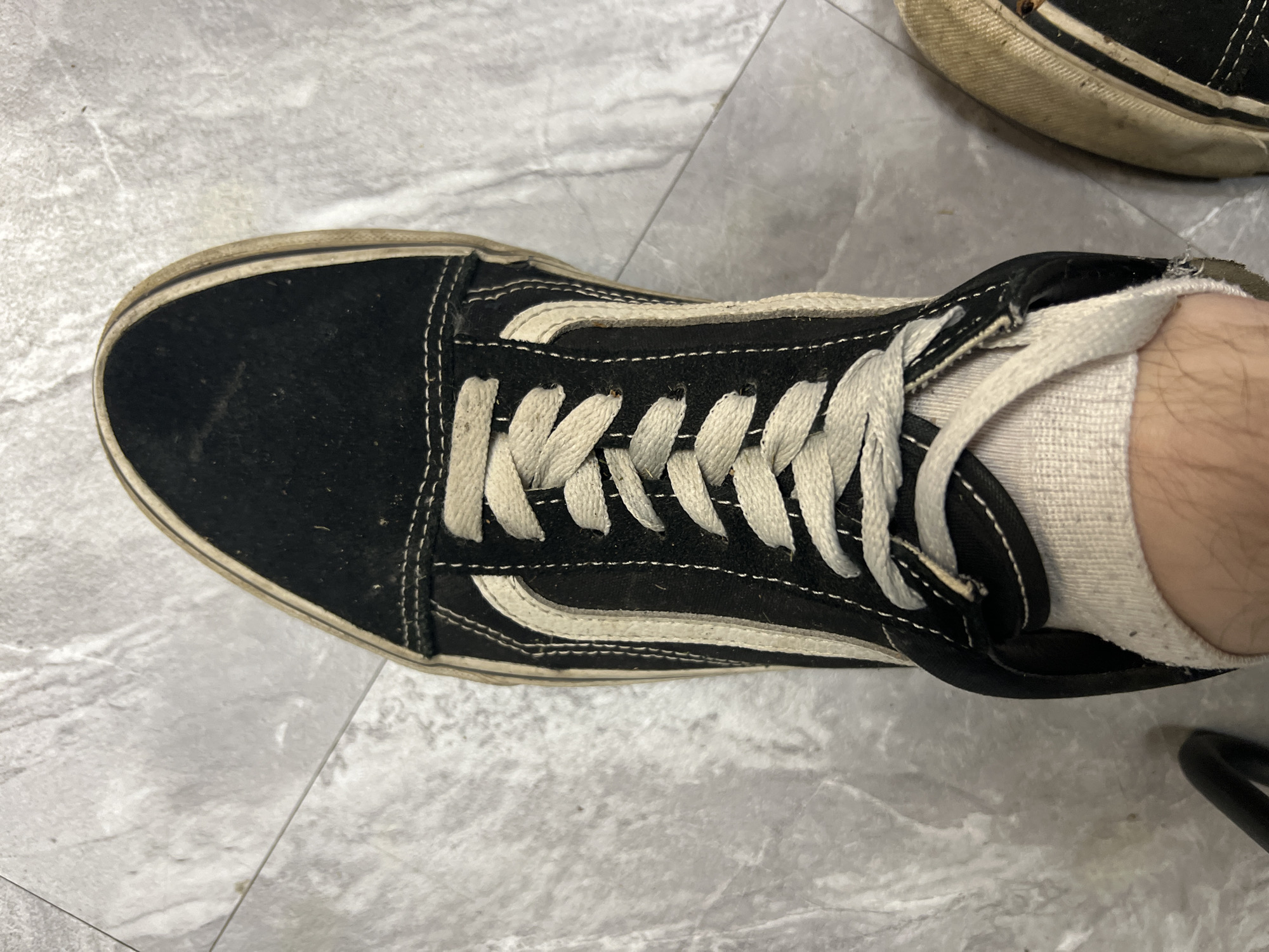A high-angle, close-up shot shows a black Vans Old Skool sneaker with white laces and a white stripe on a textured gray tiled floor. The shoe is worn and slightly dirty, with visible wear on the sole and fabric. A portion of a white sock is visible around the ankle area, and the leg of the wearer is also visible. Another similar shoe is partially visible on the right side of the frame. The environment appears to be indoors, possibly a home or a public space with tiled flooring. There is no indication of the shoe's location in Ленинский, Russia.