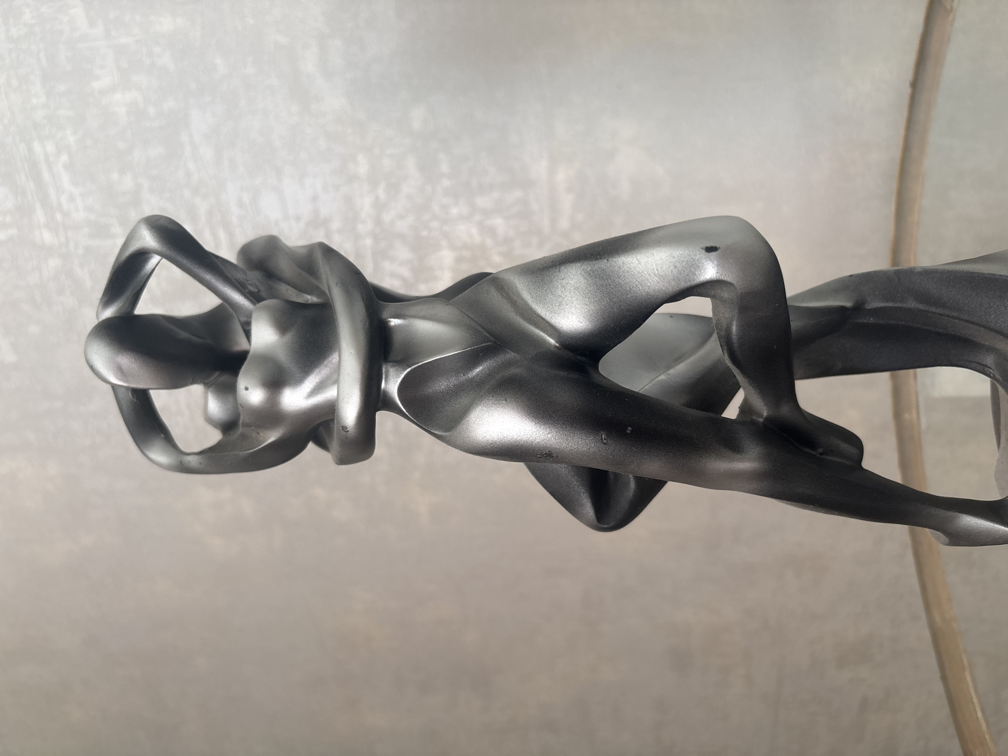 The image shows a close-up of a metallic, abstract sculpture of two figures embracing. The sculpture is dark gray with a slight sheen, and the figures are stylized with smooth, flowing lines, suggesting movement and intimacy. One figure appears to be behind the other, with arms wrapped around the torso. The background is a textured, light-colored wall, possibly concrete or stucco, with subtle variations in tone and possibly some subtle horizontal lines. The lighting is soft, creating highlights and shadows that emphasize the sculpture's form. The base of the sculpture is not fully visible, but a portion of a reflective surface, possibly glass or polished metal, is present at the bottom. There are no people, animals, or any other identifiable objects present in the image. The image is a still photograph of a decorative art piece. The location in Krasnodar, Russia, is not depicted or suggested in the visual information provided.
