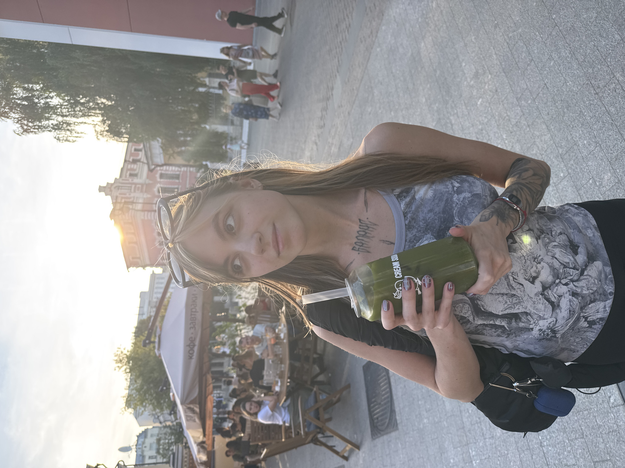 A young woman with long, light brown hair is standing on a cobbled street in Moscow, Russia, holding a green beverage in a can with a straw. She is wearing a gray tank top with a black and white graphic print and sunglasses pushed up onto her hair. A black backpack is slung over her left shoulder, with two small pouches attached to its strap. Tattoos are visible on her arms and chest.
In the background, people are seated at tables outside a cafe with a large umbrella providing shade. The cafe sign reads "Кофе. Завтраки." (Coffee. Breakfasts.). Further in the background, several people are walking down the street. Buildings with a distinct architectural style are visible, bathed in the warm light of the setting sun. The overall atmosphere suggests an outdoor dining area in a city setting during the evening.