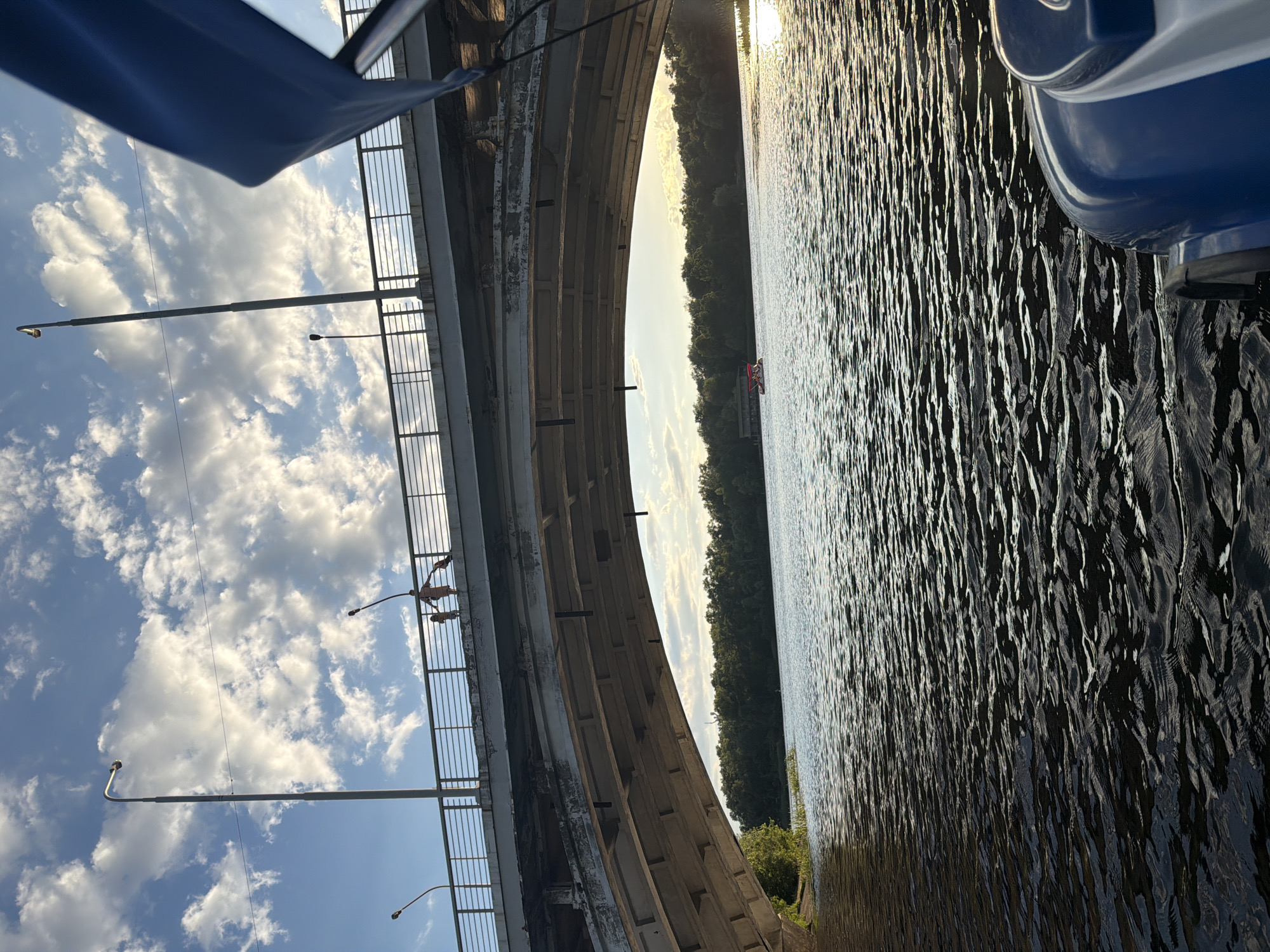 The image depicts a scene from under a concrete bridge, looking out towards a body of water and a treeline. The perspective is from a boat, as evidenced by the blue object in the upper right corner, likely part of the boat's structure.
The main subjects are the bridge, the water, and the sky. The bridge is a large, curving concrete structure with visible support beams. A railing runs along the top of the bridge, and two streetlights are visible on the left side, attached to poles extending from the bridge. A thin wire stretches across the sky, possibly an electrical line.
On the bridge, two figures are visible standing near the railing. They appear small in comparison to the vastness of the bridge and the landscape.
The water is dark and rippling, reflecting the sunlight which creates streaks of light on its surface. This suggests it is either late afternoon or early morning. The sky is blue with scattered white clouds. The treeline on the far bank is dense and green, with a few patches of lighter sky visible beyond it. A small red boat or raft can be seen in the distance on the water.
The scene appears tranquil, with the natural elements of water, sky, and trees contrasting with the man-made structure of the bridge. The lighting suggests a pleasant weather condition. There is no visible text in the image.