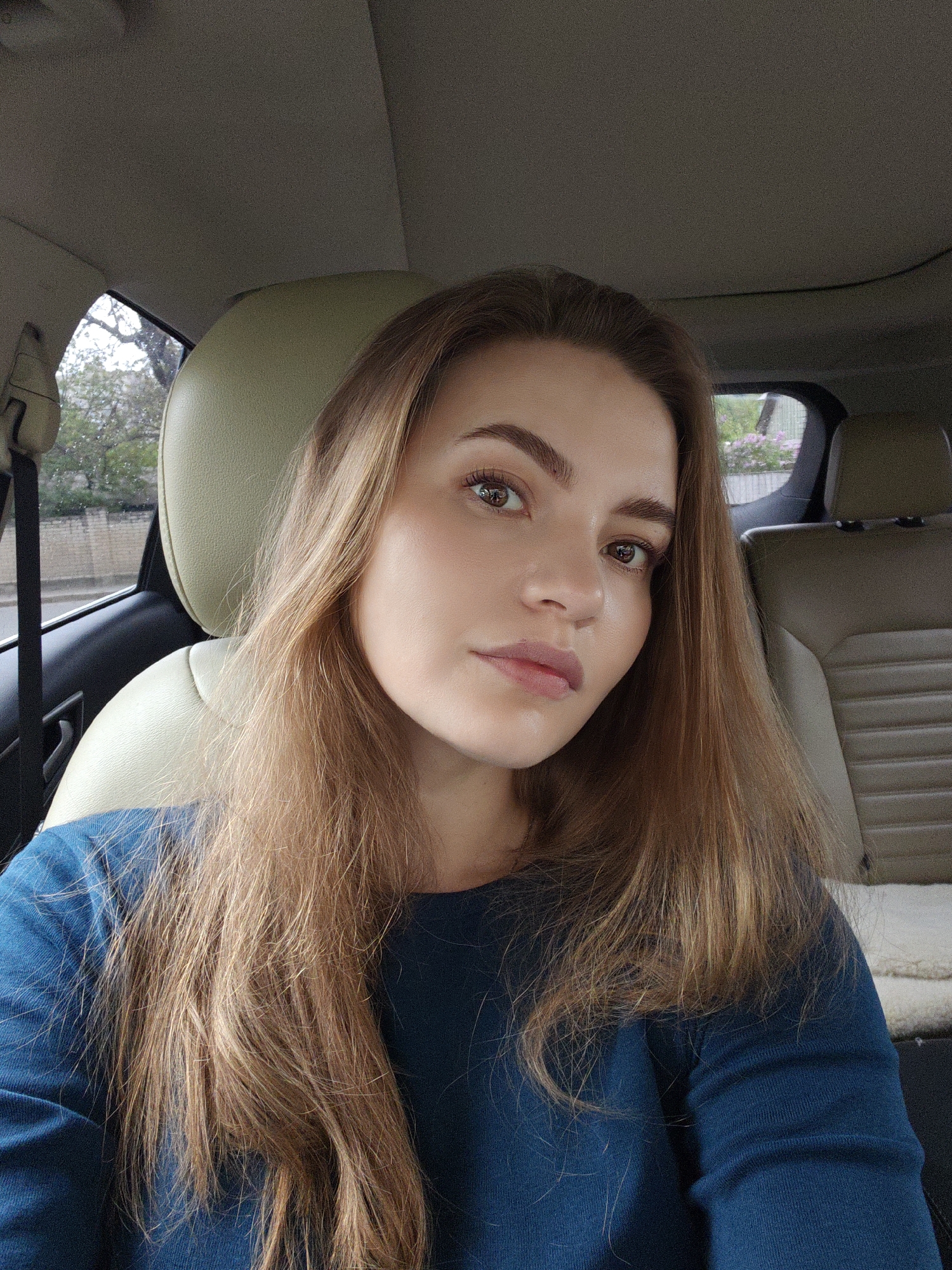 The image shows a woman in the front seat of a car. She has light brown hair, fair skin, and brown eyes. She is wearing a teal long-sleeved shirt. The car's interior is beige leather. The background shows trees and a building through the window. The location cannot be definitively determined as Vinnytsia, Ukraine from the image alone.