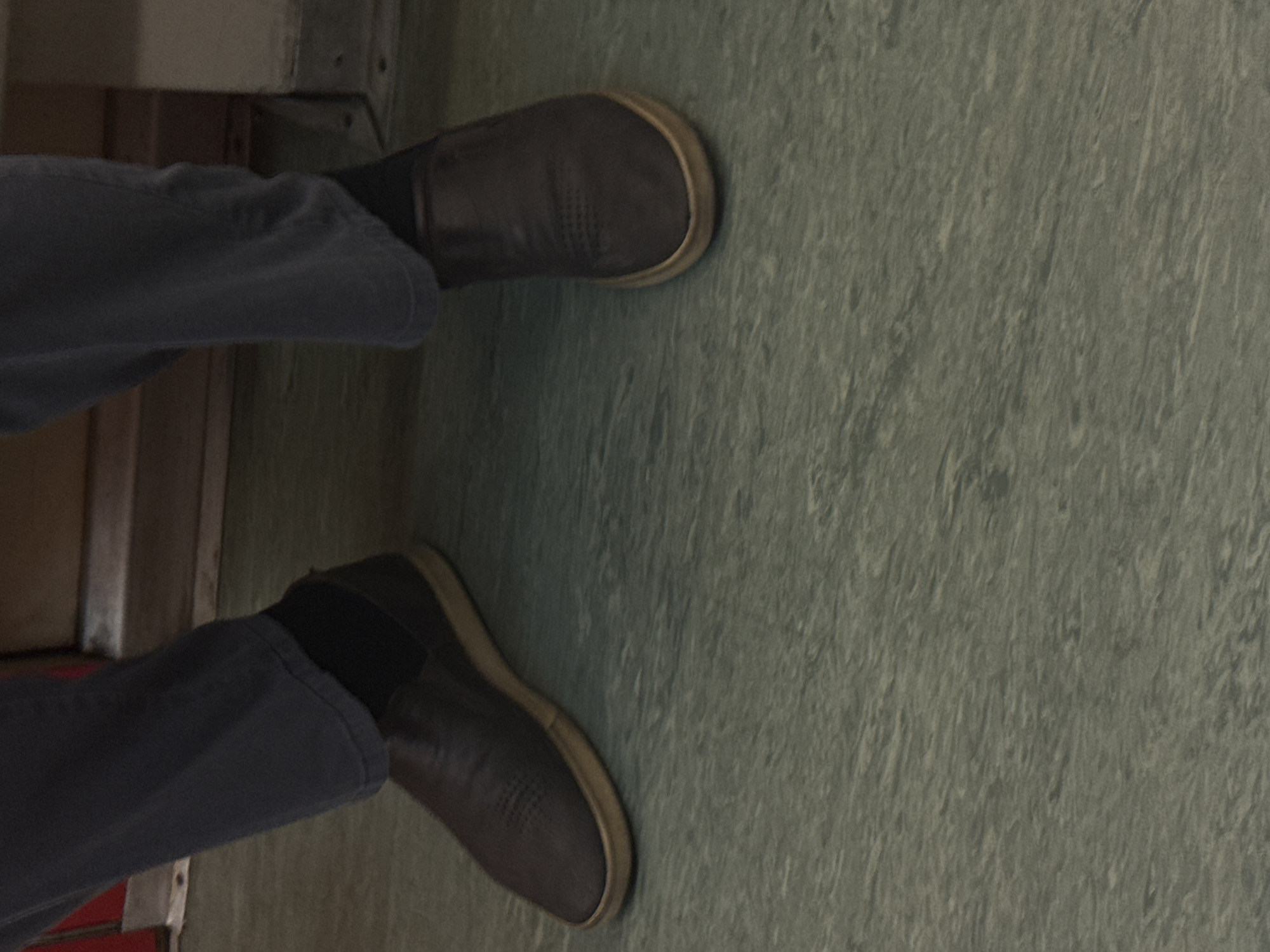 The image shows a pair of legs in jeans and slip-on shoes, likely inside a public transport vehicle. The floor has a green, textured surface. The setting could plausibly be a train or bus in a city, but specific details to confirm it is Moscow, Russia, are absent.
