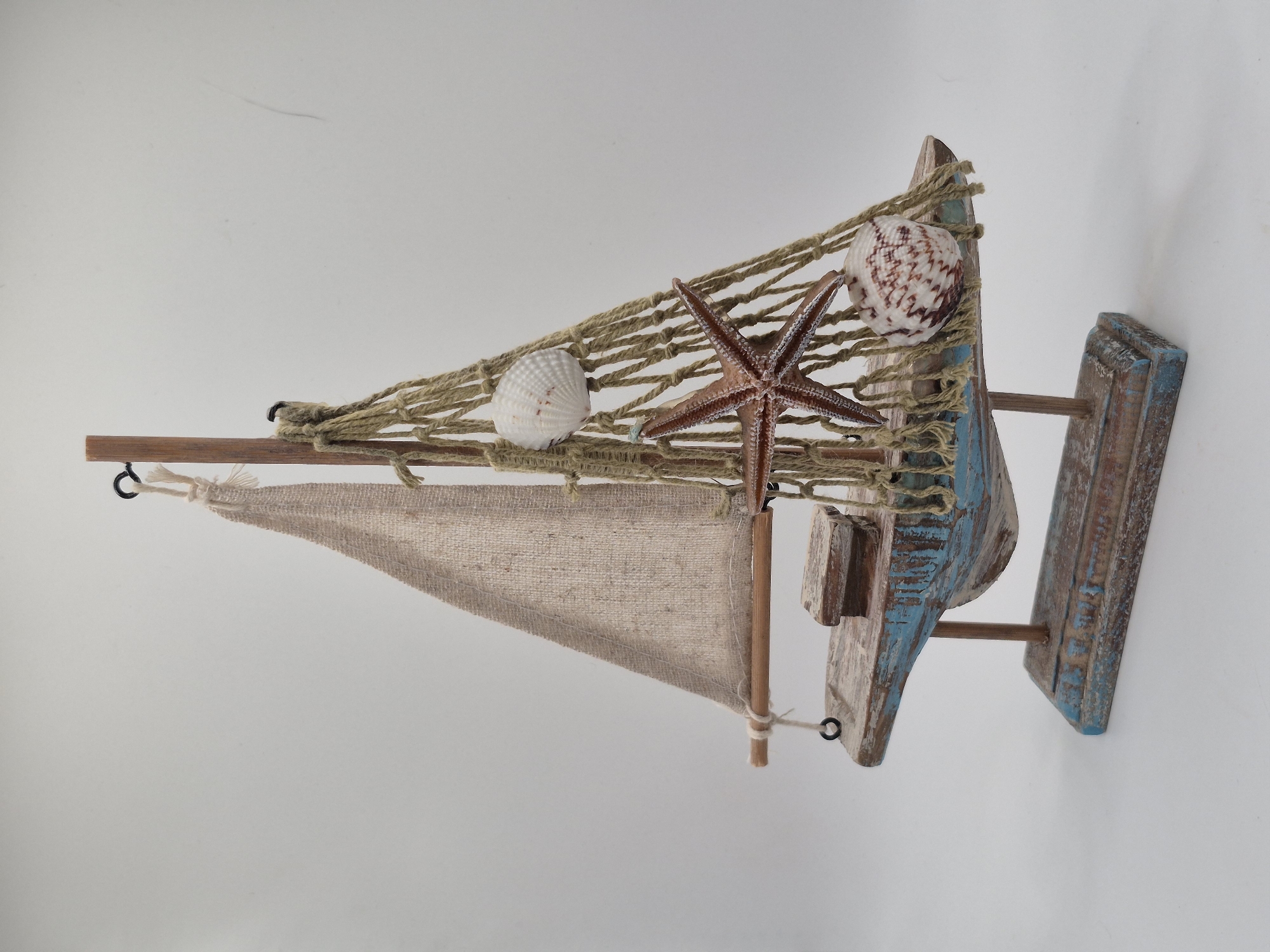 A decorative wooden sailboat model is displayed against a plain, light-colored background. The sailboat has a weathered appearance with blue and brown painted accents on its hull and base. It features a tan fabric sail, a wooden mast, and netting draped around it. Three nautical embellishments are attached to the netting: a brown starfish and two seashells, one white and one reddish-brown and white. The model is supported by two wooden rods that connect to a rectangular wooden base. The lighting is even, casting soft shadows. There are no people, animals, or any signs of activity. The overall setting appears to be a studio or display area, not a location within Schwäbisch Gmünd, Germany.