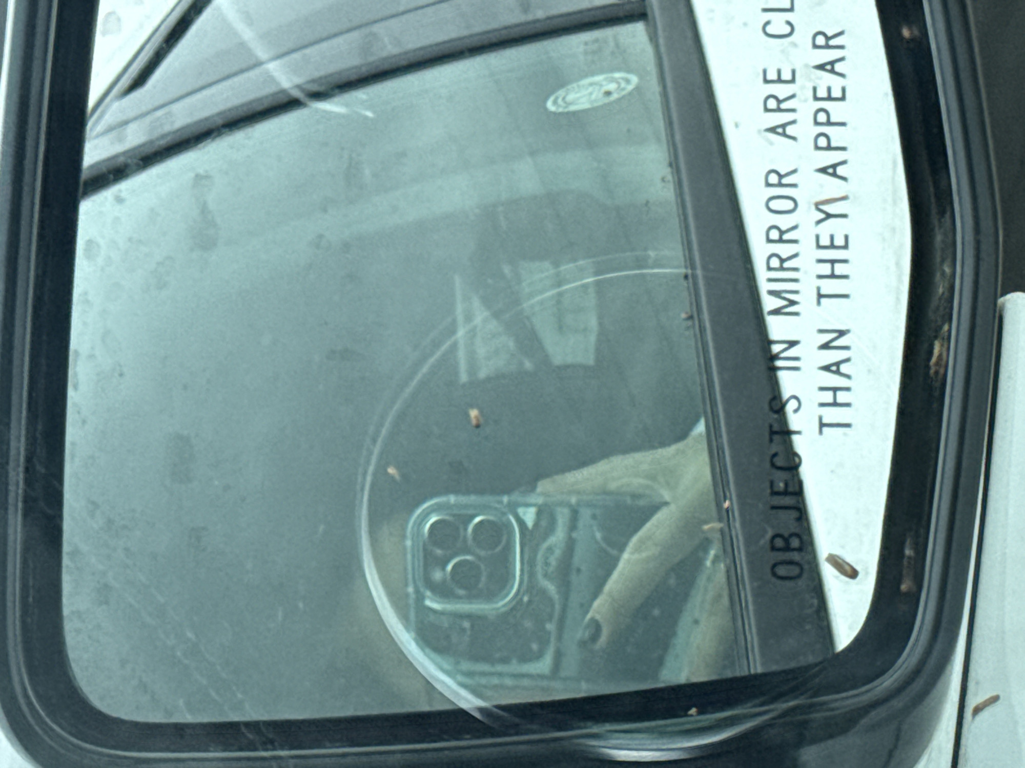 The image shows a car's side mirror with the inscription "OBJECTS IN MIRROR ARE CLOSER THAN THEY APPEAR." Reflected in the mirror is a hand holding an iPhone with a clear case, capturing the reflection. The mirror itself appears slightly dirty. The location is New Braunfels, United States of America.