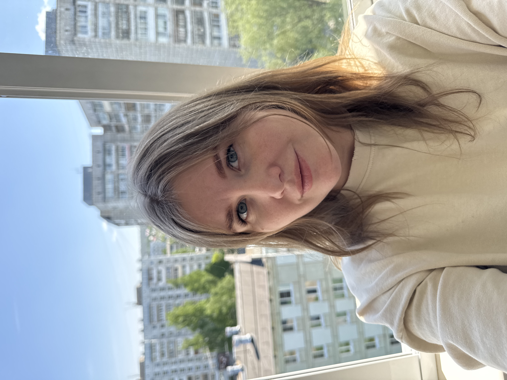 A selfie of a woman with light brown hair and blue eyes, wearing a cream-colored sweatshirt, is positioned in front of a window with a view of a city. The city scene includes tall apartment buildings, greenery, and a clear blue sky. Based on the architectural style of the buildings, the location is likely Chaikovsky, Russia. The woman is looking at the camera, with a slight smile.