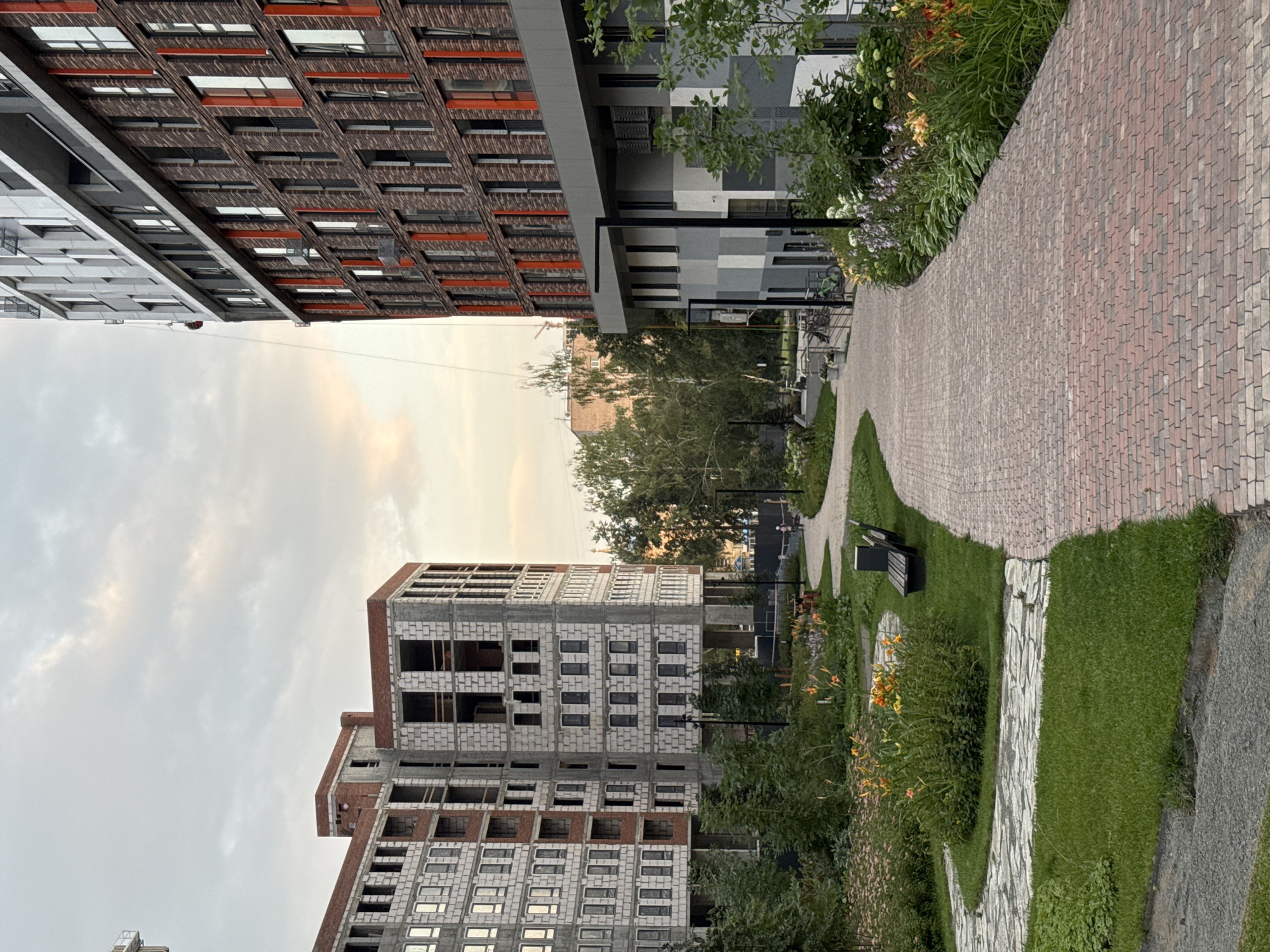 The image depicts a courtyard area in a modern residential complex under construction. Two multi-story buildings are visible; one appears to be nearing completion with brickwork and windows installed, while the other is in an earlier stage of construction with exposed concrete blocks and unfinished window openings. A paved walkway made of red and grey bricks winds through the courtyard, bordered by green grass, flowering plants, and trees. Benches are placed along the path. The sky is overcast with hints of sunset colors. The overall scene suggests an urban environment with ongoing development. This scene is located in the city of Izhevsk, Russia.