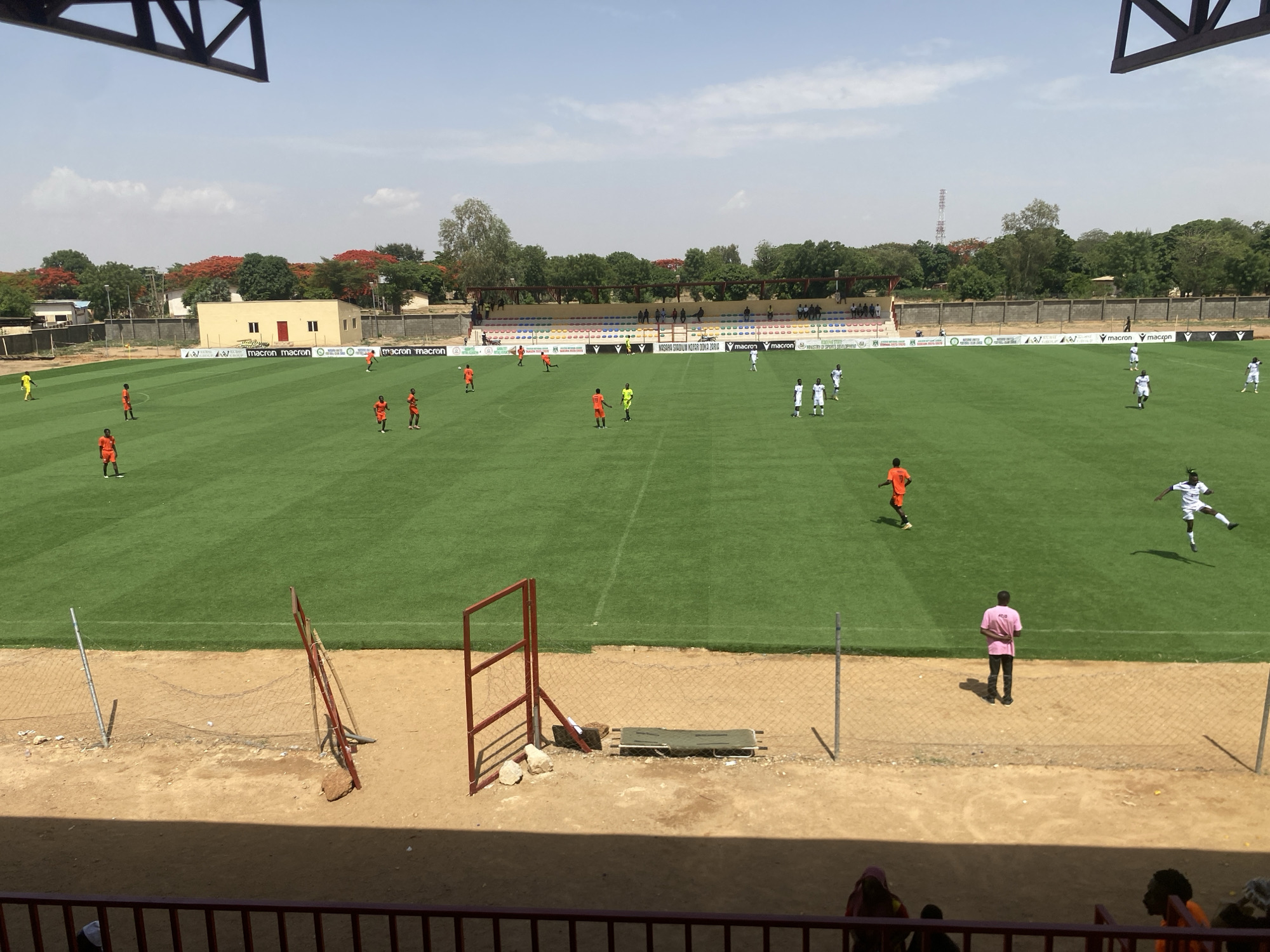 The image depicts a soccer match in progress at the Ahmadu Bello Stadium in Tudun Wada, Nigeria. Players in orange and white uniforms are actively engaged on a well-maintained green pitch. Several individuals are in the stands, with one person in a pink shirt standing near the edge of the field. The stadium has a small stand in the background and is bordered by trees and buildings. A communications tower is visible in the distance. A metal fence separates the stands from the field.