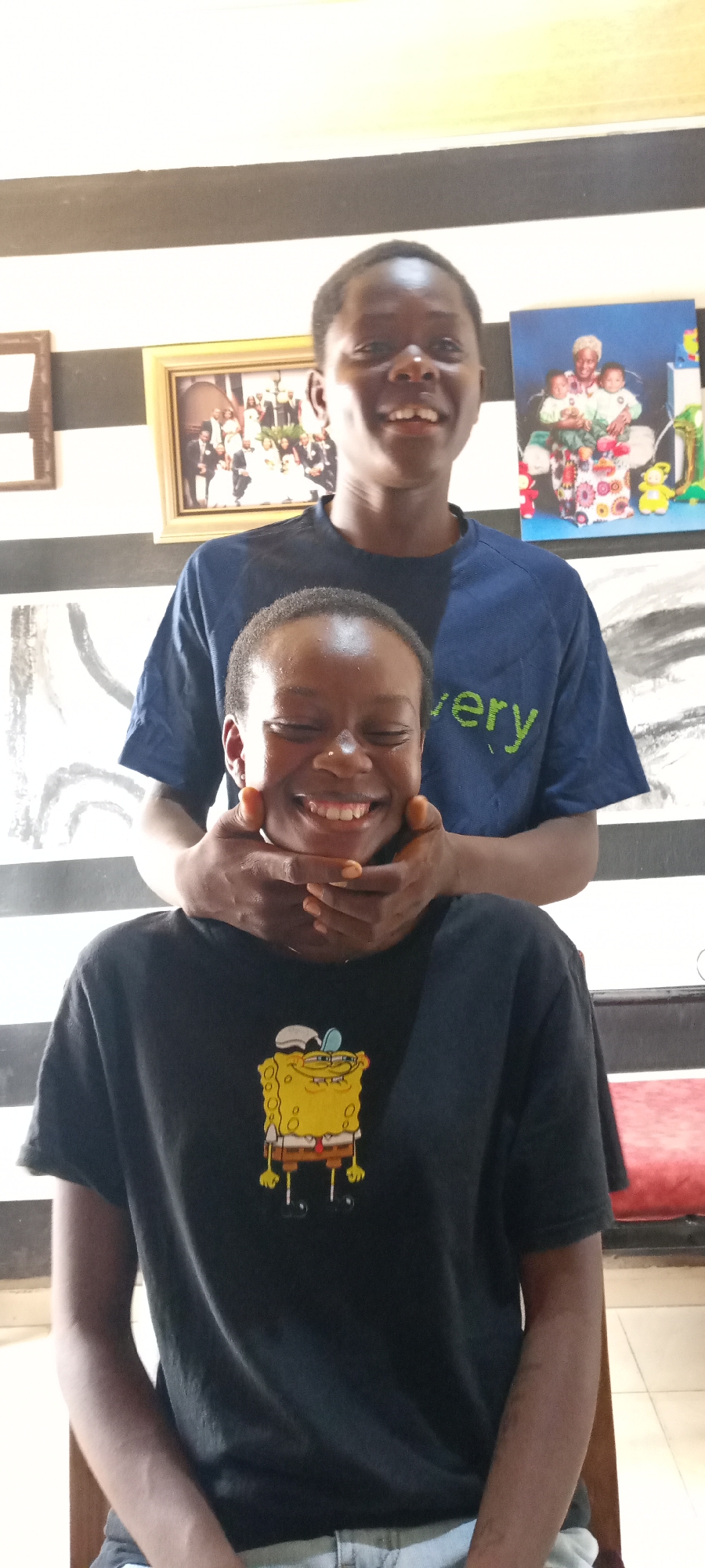 Two dark-skinned individuals are pictured indoors, one seated and one standing behind. The seated individual wears a black T-shirt with a SpongeBob SquarePants graphic and has their eyes closed, smiling. The standing individual wears a blue T-shirt and has their hands around the seated person's neck and chin. The location features a black and white striped wall, with framed pictures hanging on it. The setting suggests an indoor environment, potentially a home or studio in Jos, Nigeria.