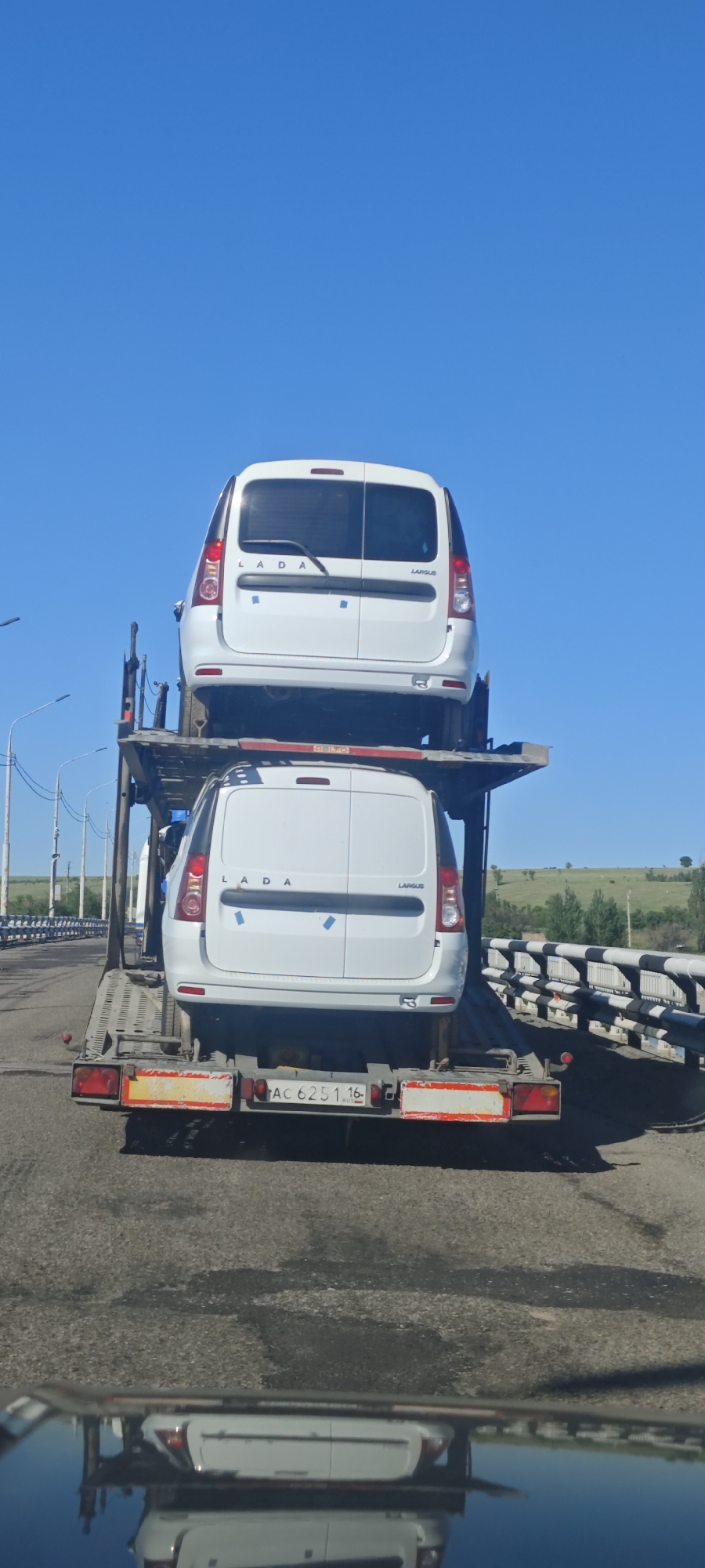 The image shows a car carrier truck transporting two white LADA Largus vans on a bridge in Ferma Sovkhoz Balykleyskiy, Russia. The truck's license plate reads "AC 6251 16 RUS." The sky is clear and blue. Guardrails line the side of the bridge, with fields and trees visible in the background. The lower part of the frame shows the reflection of the scene in the car mirror.