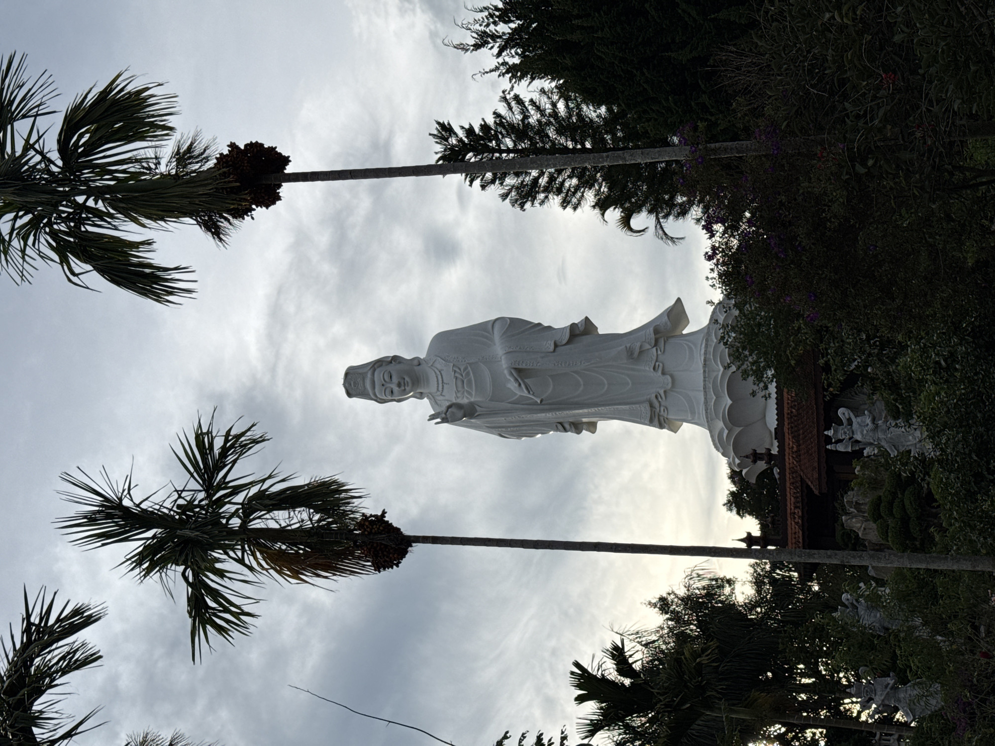 A vertically oriented shot depicts a white statue of a robed figure, likely a Buddhist deity, standing atop a multi-tiered base, framed by tall palm trees and dense foliage. The sky is overcast with grey and white clouds. The statue is situated within a garden setting, with other smaller statues partially visible among the trees. The scene is located in Thôn 5, Vietnam.