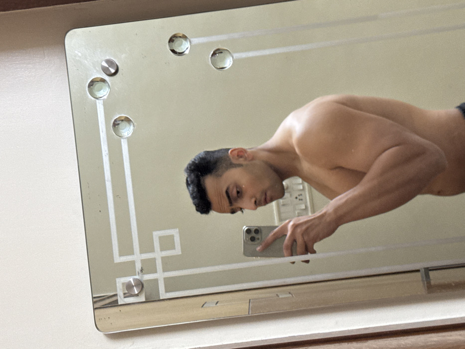 A muscular man is reflected in a framed, rectangular mirror with decorative embellishments. The mirror features inlaid geometric designs and round, jewel-like accents along its border. The man is shirtless, holding a smartphone to capture the reflection. He is positioned close to the mirror, and his face and upper torso are visible. The background appears to be a lightly colored wall, and an electrical outlet is also reflected in the mirror.