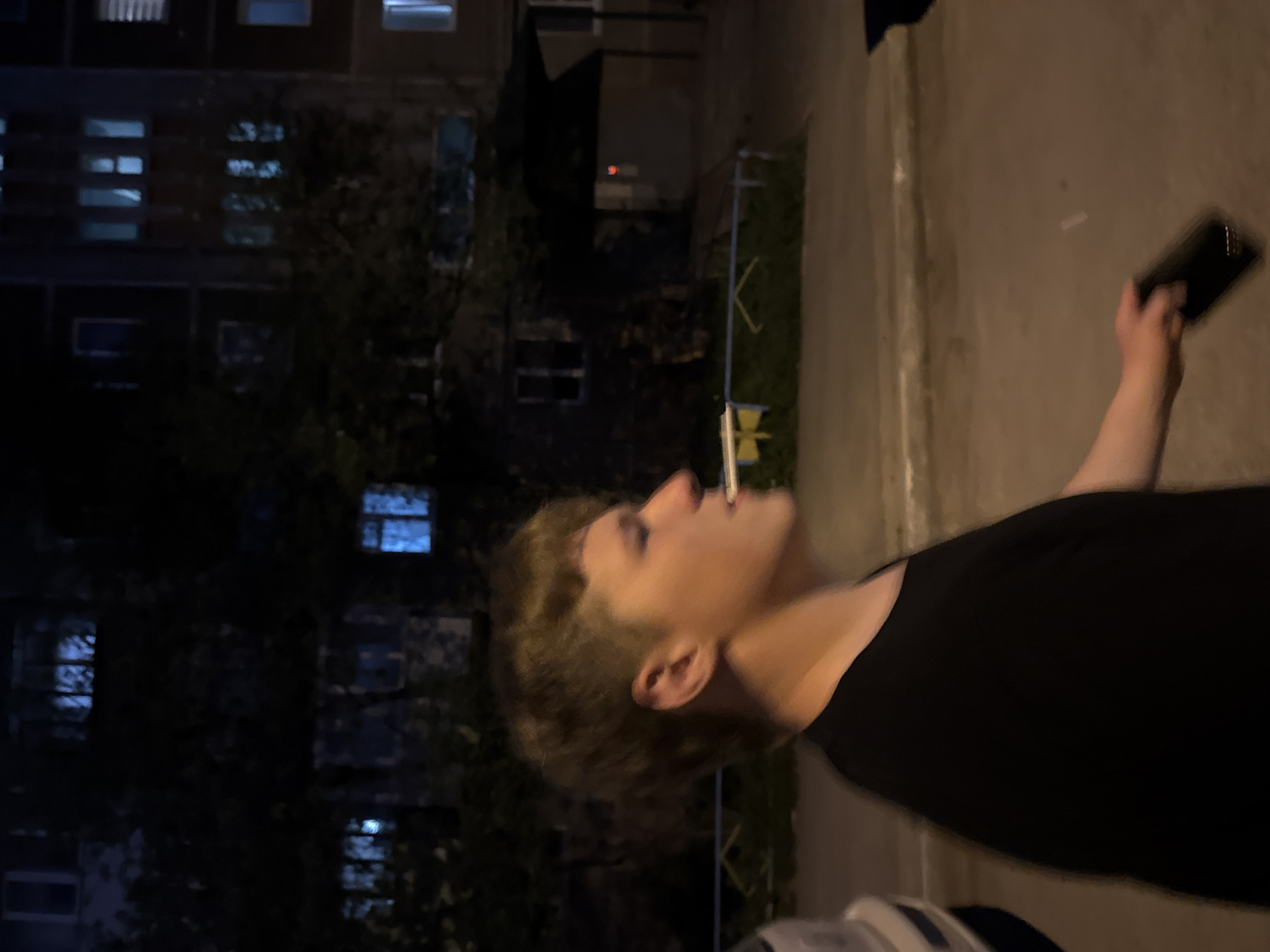 A person stands outside an apartment building in what is likely Perm, Russia, based on location and the architectural styles common in Russian urban areas. The person holds a mobile phone in one hand and has a cigarette in their mouth. The apartment building in the background has numerous windows, some with lights on, suggesting it's nighttime. The area appears to be a courtyard or walkway near the building. The scene is illuminated by artificial lighting.