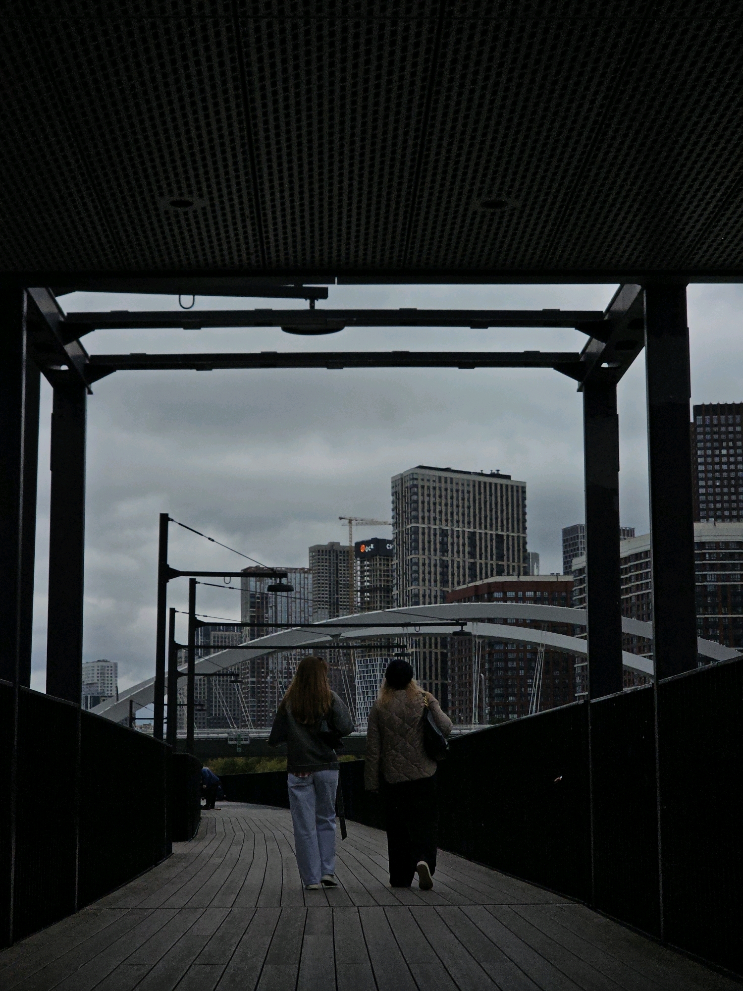 Two women walk away from the camera on a wooden walkway. The woman on the left has long, blond hair and wears a grey hooded sweatshirt and light blue jeans. The woman on the right wears a quilted beige jacket, dark pants, and a black beret. They are walking towards a city skyline with various modern apartment buildings. The sky is overcast. The walkway appears to be part of a modern pedestrian bridge or elevated pathway within a contemporary urban development. The architecture suggests a location in a developed city, possibly Moscow, Russia, given the style of the buildings which are characteristic of newer residential complexes in the capital. The overall atmosphere is muted due to the cloudy weather and the dark tones of the foreground structure.
