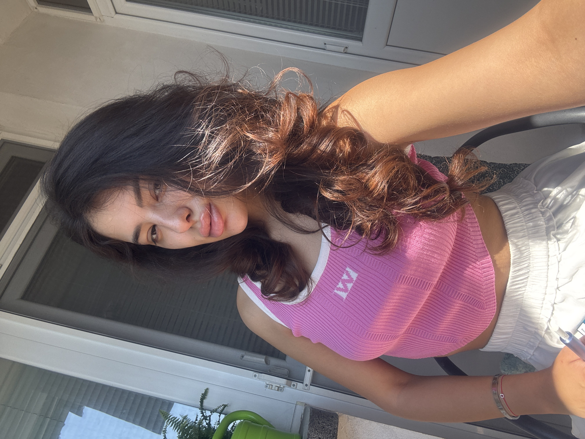 A young woman with dark, wavy hair and full lips is taking a selfie outdoors. She is wearing a pink ribbed tank top with white trim and a white textured skirt. She has on bracelets on her left wrist and is holding a blue and white object, possibly a vape, in her right hand. The background suggests a balcony or patio area, with a white door and window with blinds visible. There is also a green watering can with a fern in the upper left corner. The lighting indicates it is daytime, with sunlight casting shadows. The location is Plovdiv, Bulgaria.